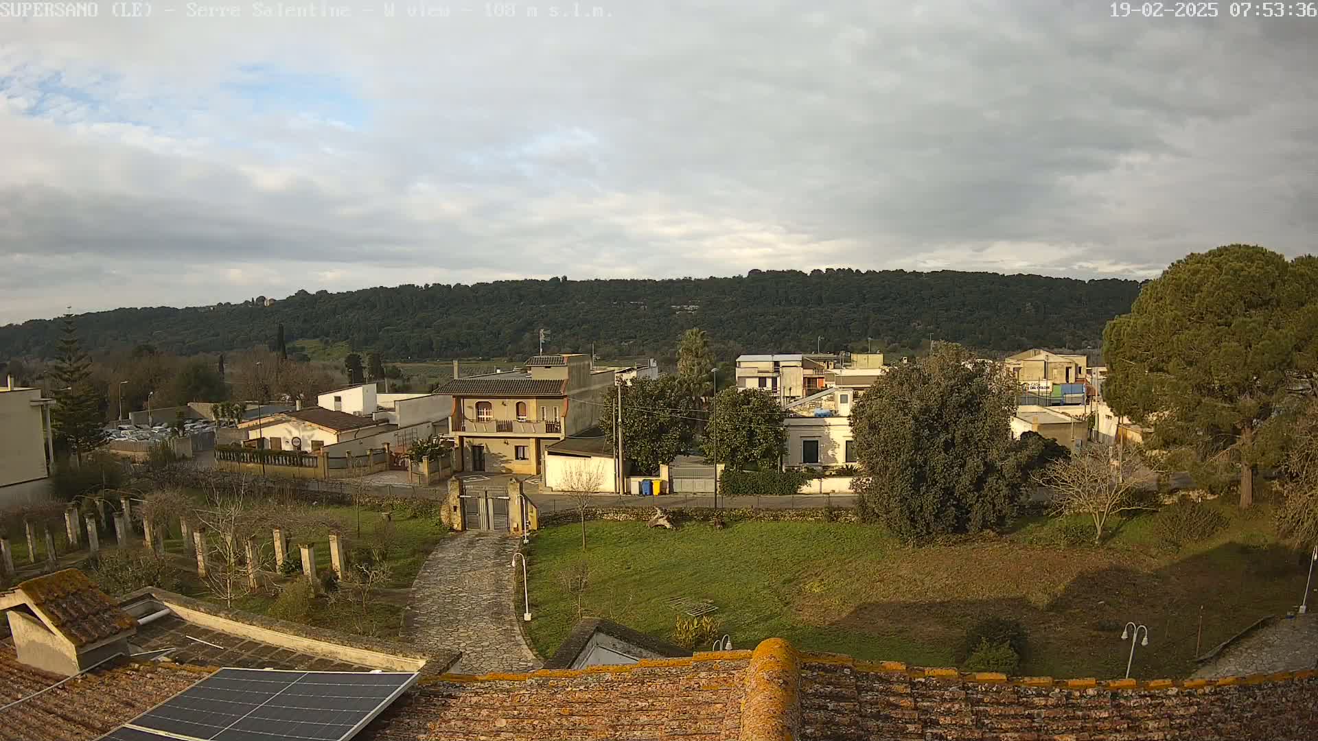 A slightly cloudy day overlooks a small town nestled below a tree-covered hill, viewed from a rooftop with a solar panel.
