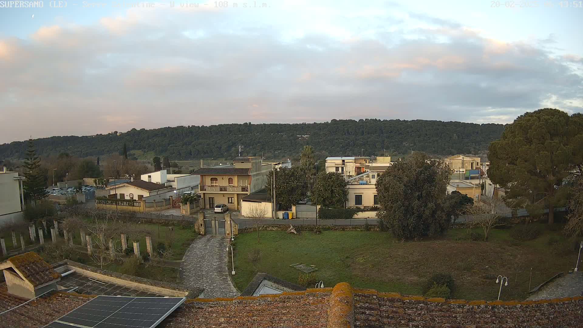 A view from a rooftop shows a residential area with low buildings, a grassy yard, and a tree-covered hill in the background under a partly cloudy sky.