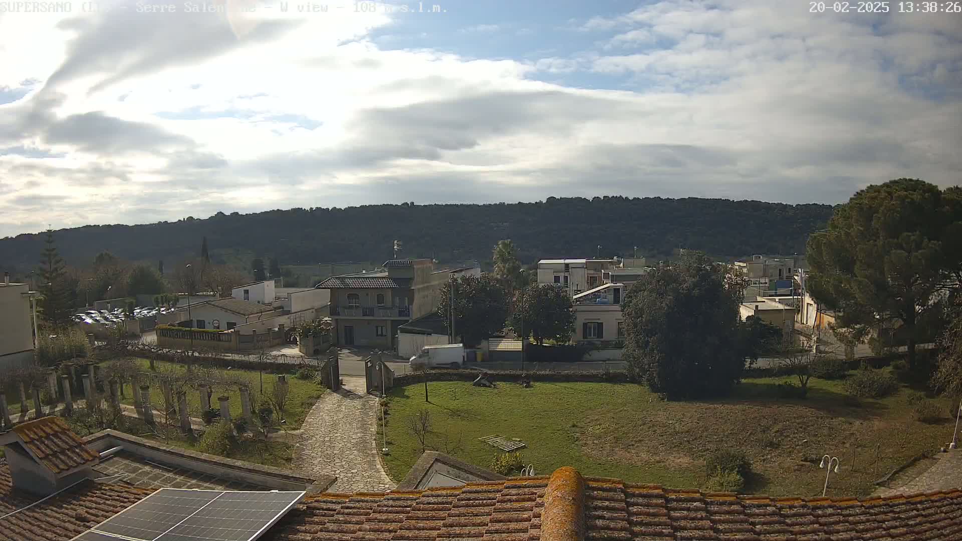 A partly cloudy sky overlooks a residential area nestled against a hillside, with a view from a rooftop featuring a solar panel.