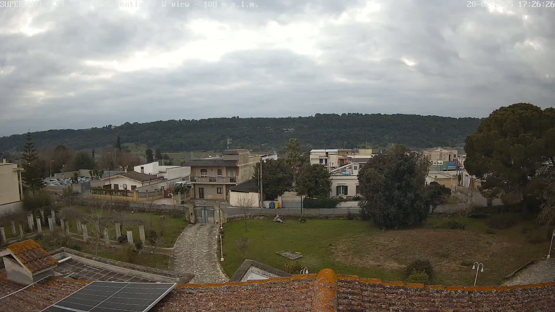 A mostly cloudy day reveals a view of a residential area nestled against a tree-covered hillside, with a stone pathway leading to a building featuring a solar panel on its roof.