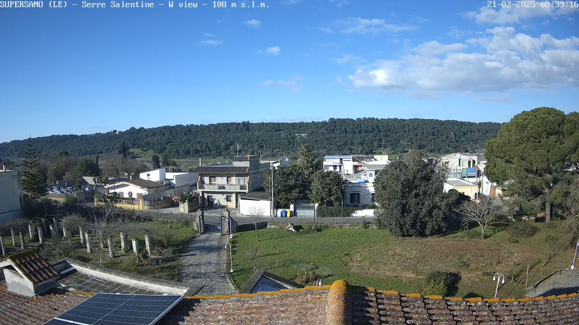 A mostly sunny day reveals a small village nestled amongst green hills, viewed from a rooftop with a solar panel.