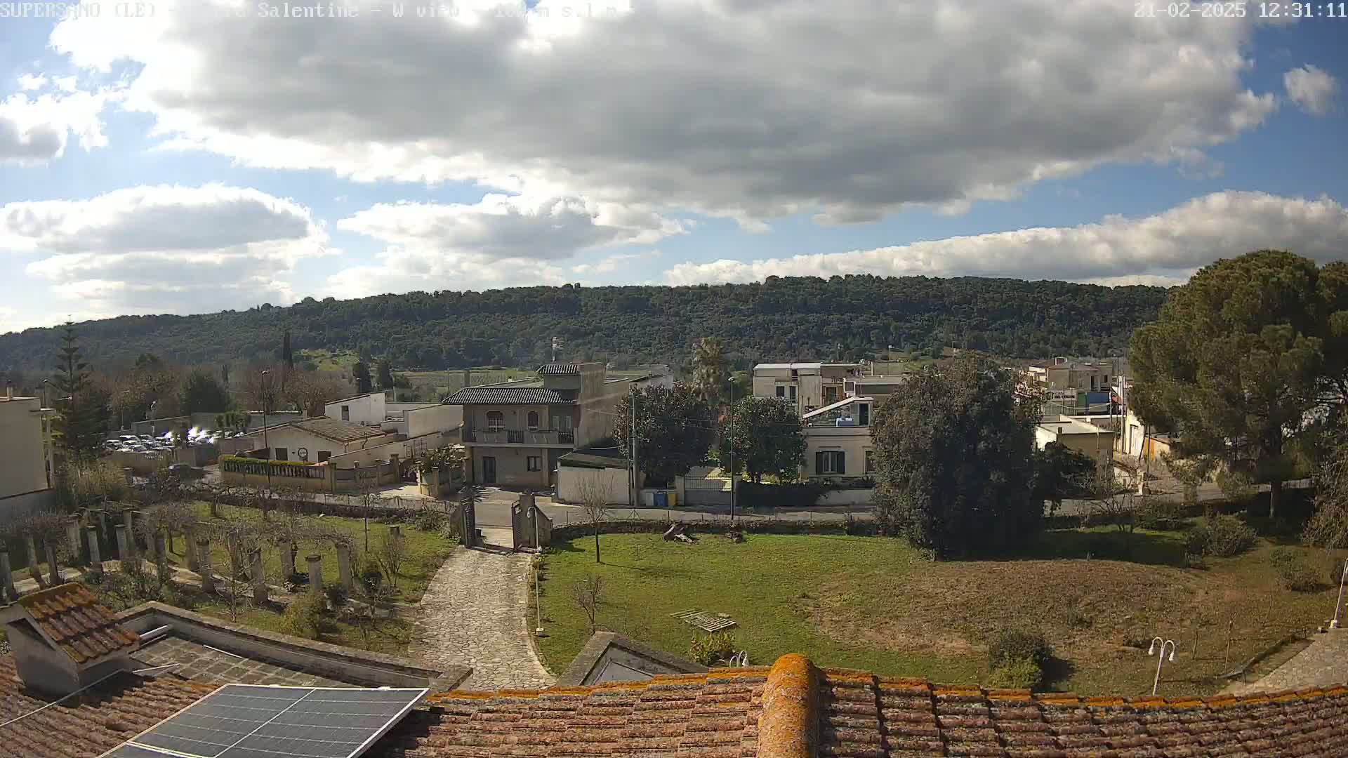 A partly sunny day reveals a small town nestled against a verdant hillside, viewed from a rooftop with a solar panel.
