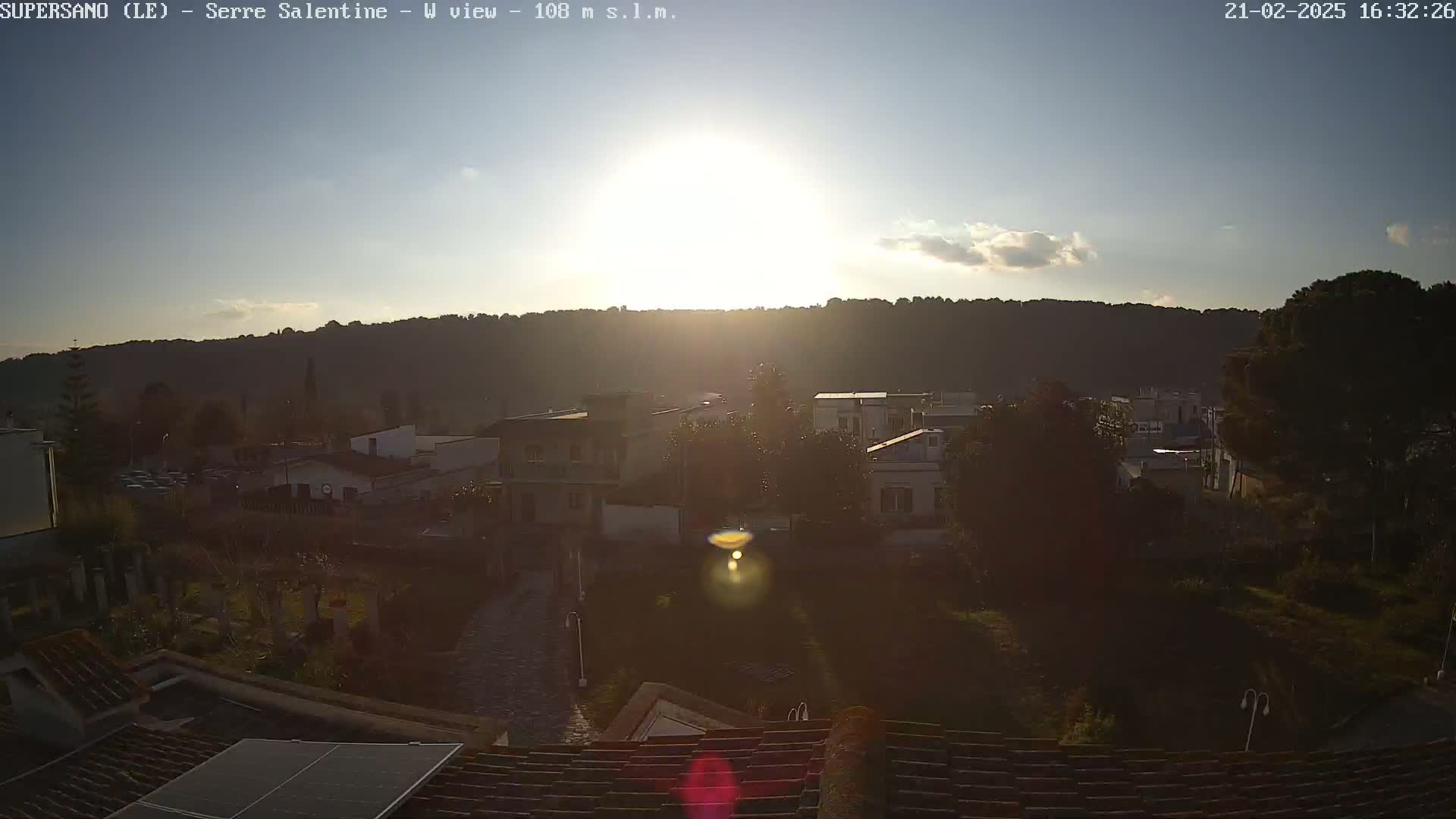 A mostly sunny sunset over a town nestled against a hillside, viewed from a rooftop with a solar panel.