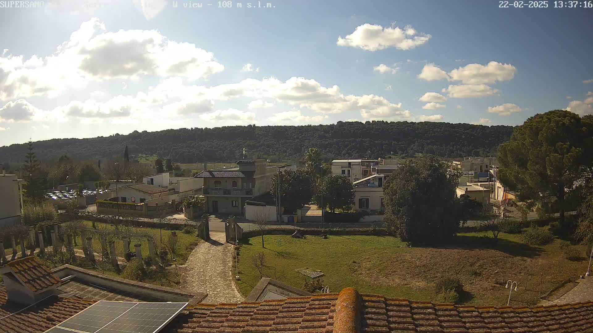 A partly sunny day reveals a view of a small town nestled against a tree-covered hill, seen from a rooftop with a solar panel.
