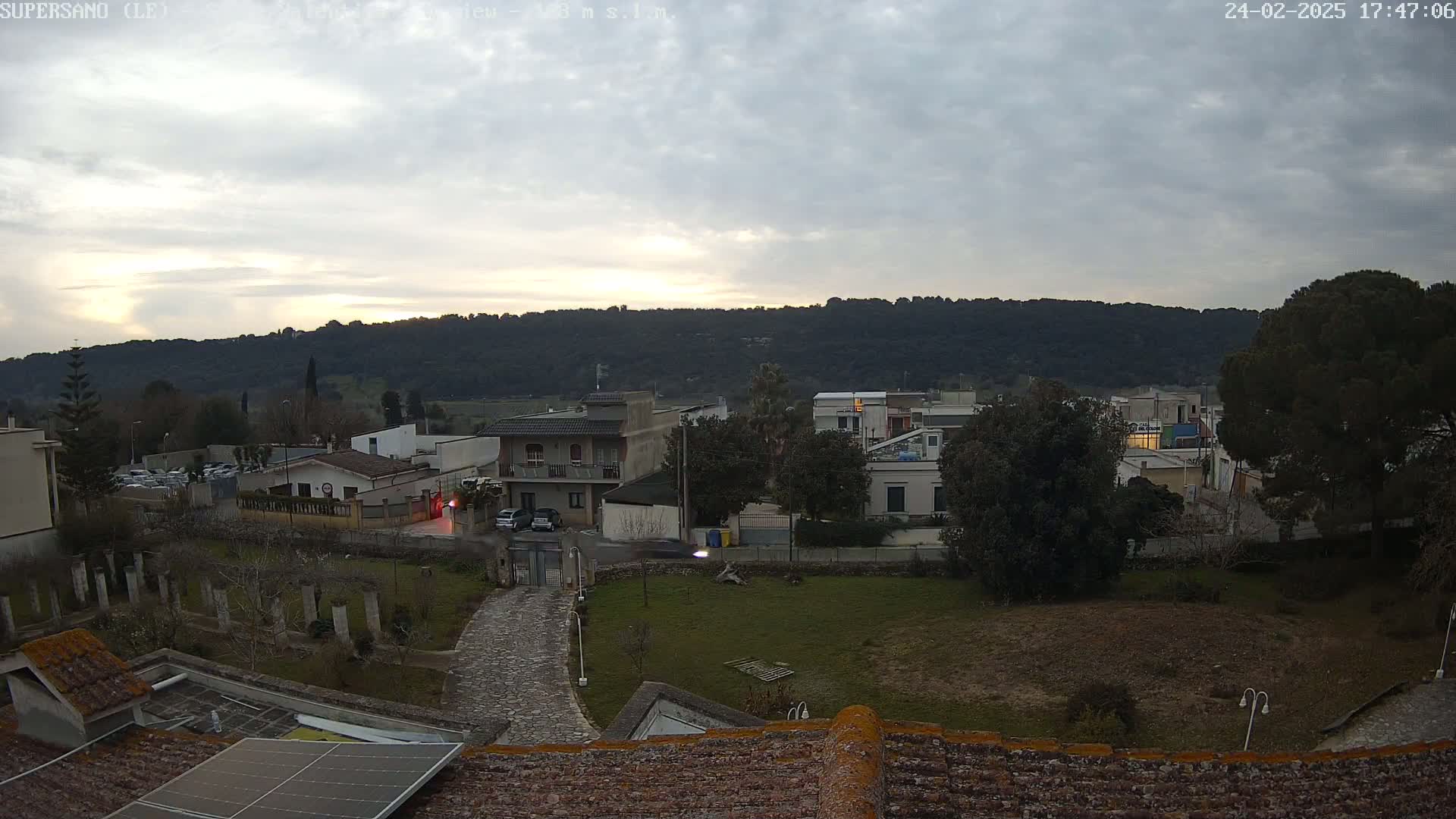 An overcast evening scene shows a residential area nestled beside a tree-covered hill, with a paved pathway leading to a house with a solar panel on the roof.