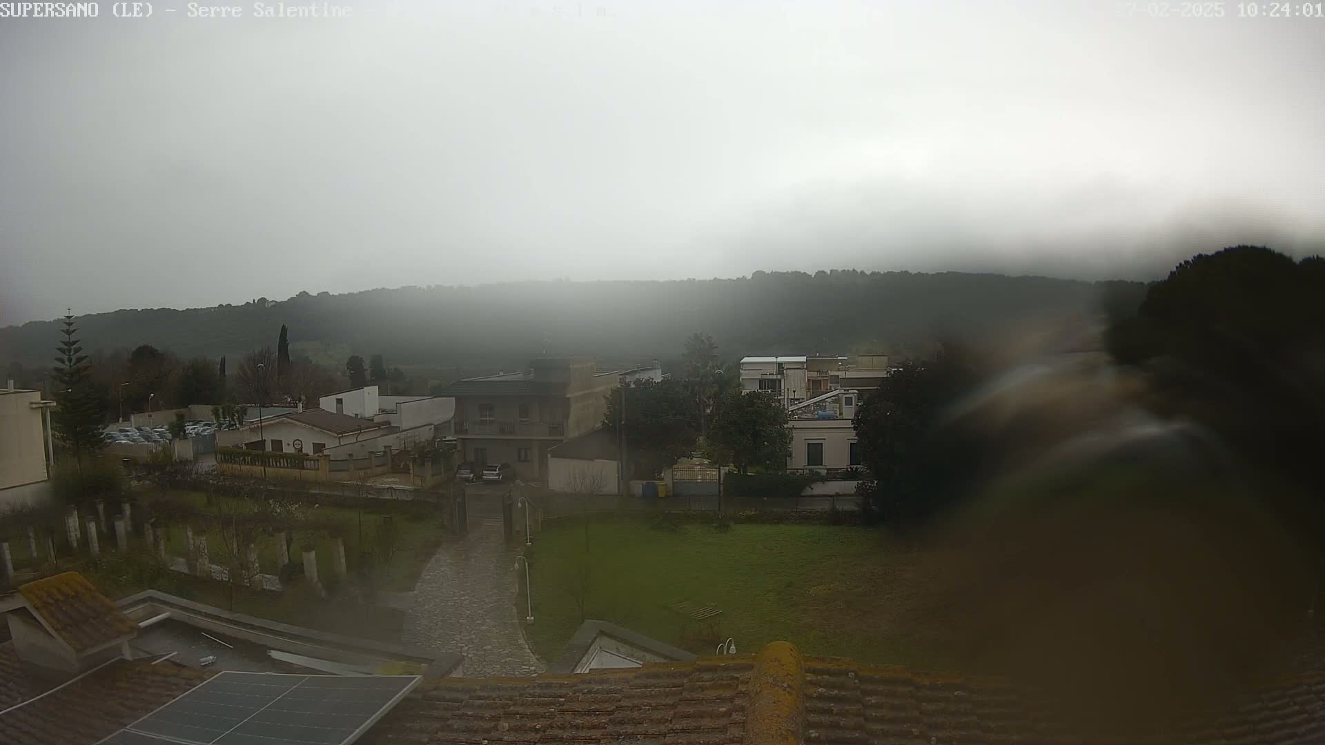A foggy, rainy view of a residential area with houses and trees, overlooking a hilly landscape.