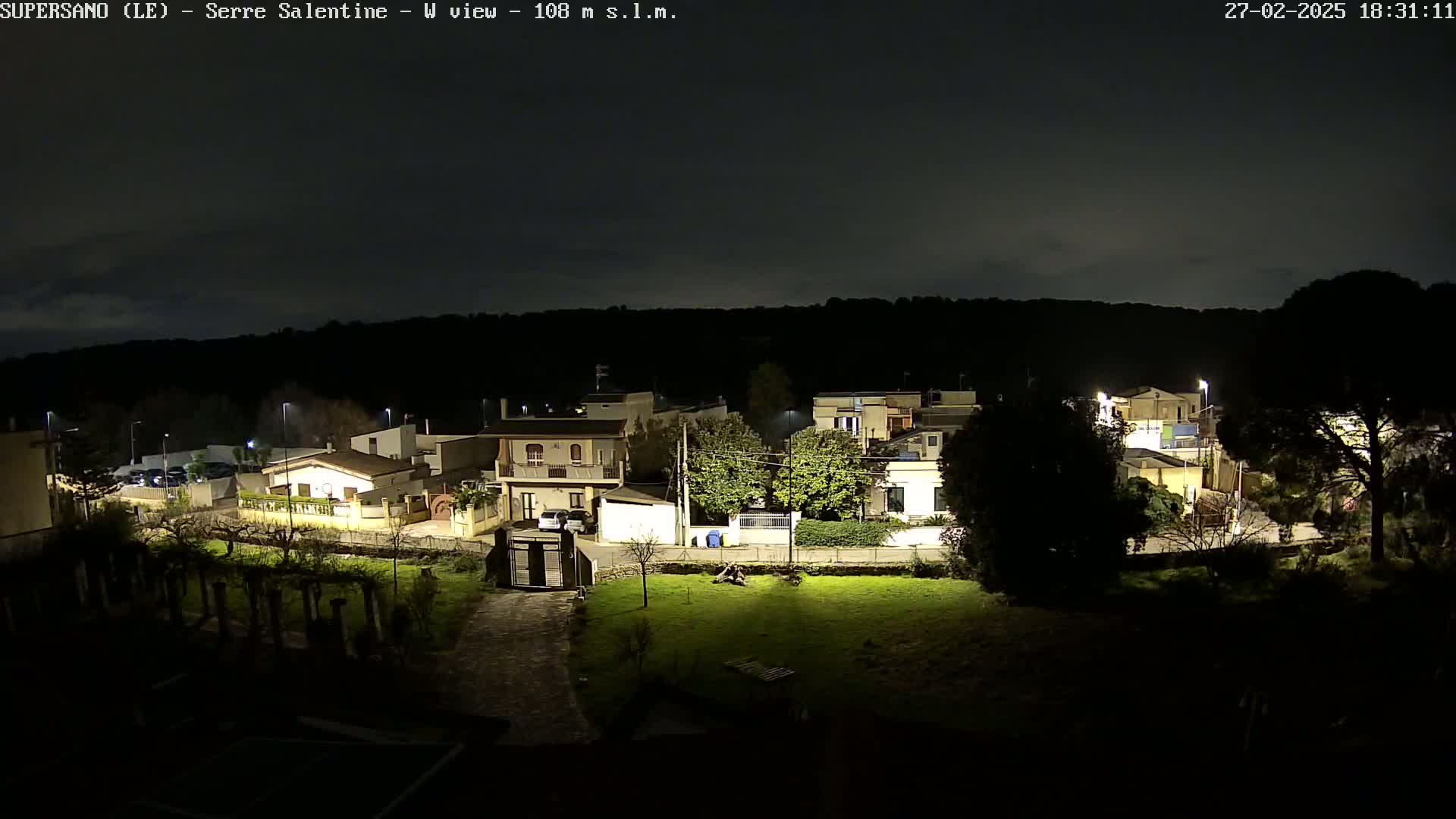 A nighttime view of a residential area with houses illuminated by streetlights, under a dark, mostly clear sky.