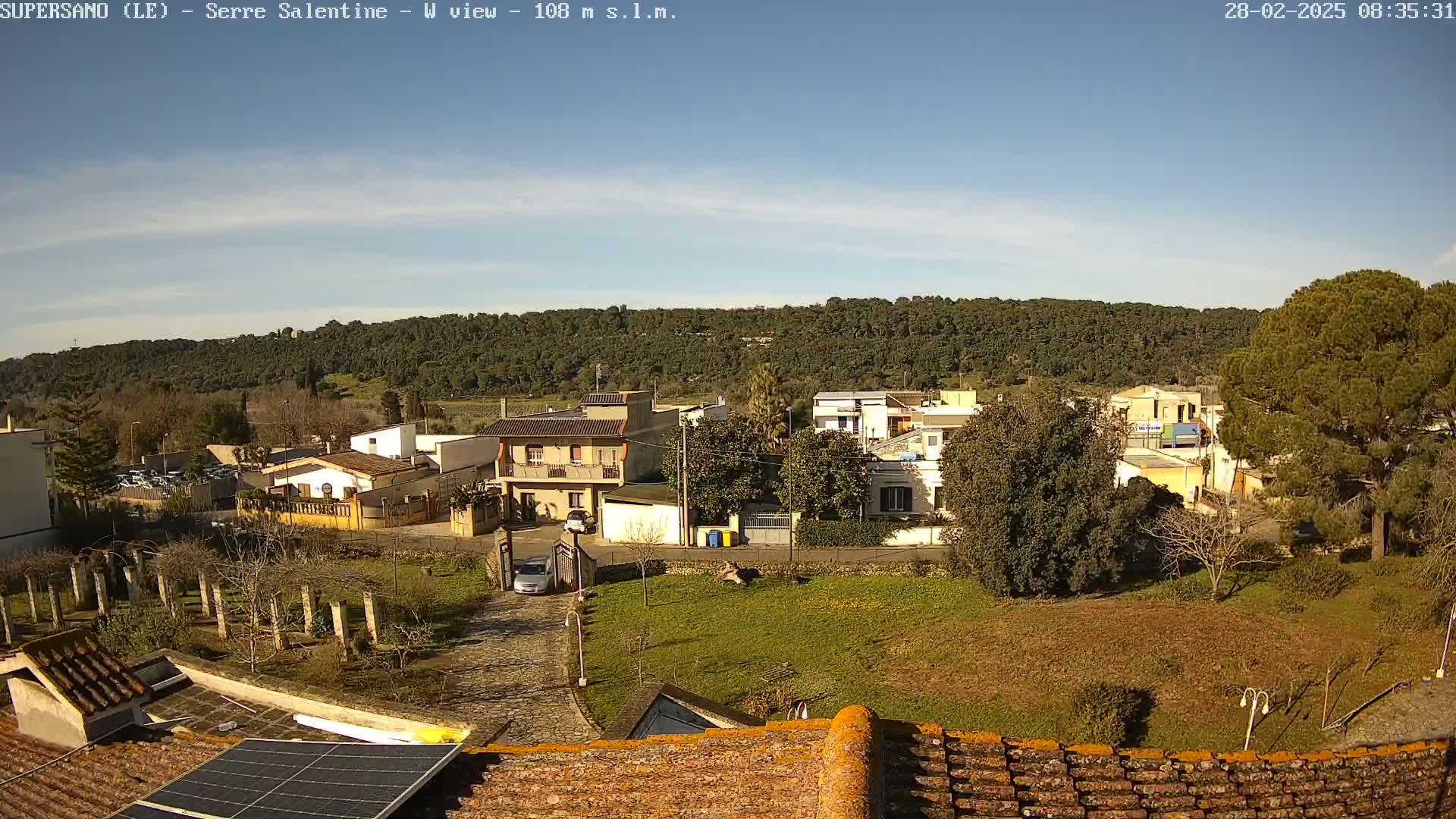 A sunny day reveals a residential area nestled against a verdant hillside, viewed from a rooftop with a solar panel.