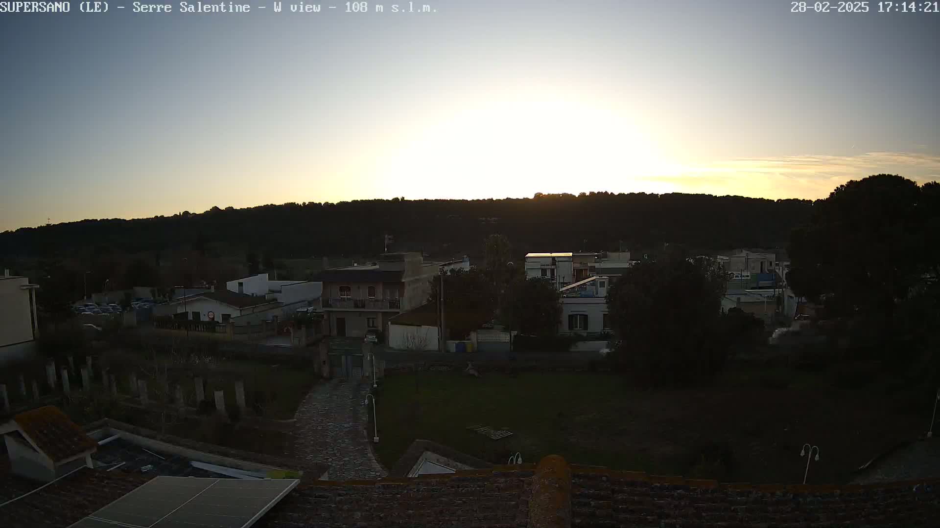 A sunset illuminates a town nestled against a hill, with the low-lying sun casting long shadows across the buildings and landscape.