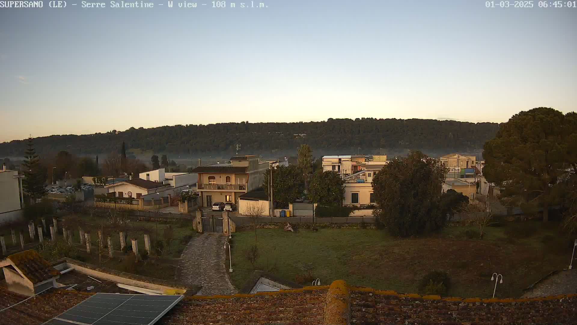 A residential area with low buildings and trees is visible, situated at the base of a hill under a clear, twilight sky.