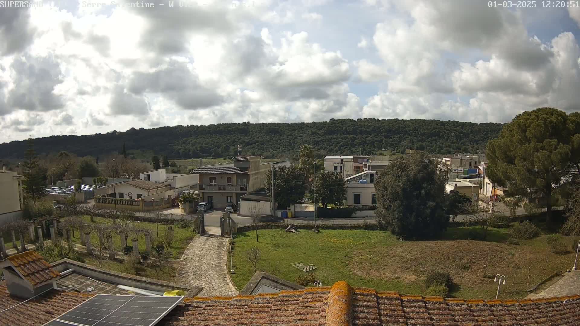 A partly cloudy day reveals a residential area nestled below a tree-covered hill, with a paved driveway leading to a gated entrance and a solar panel visible on a nearby roof.