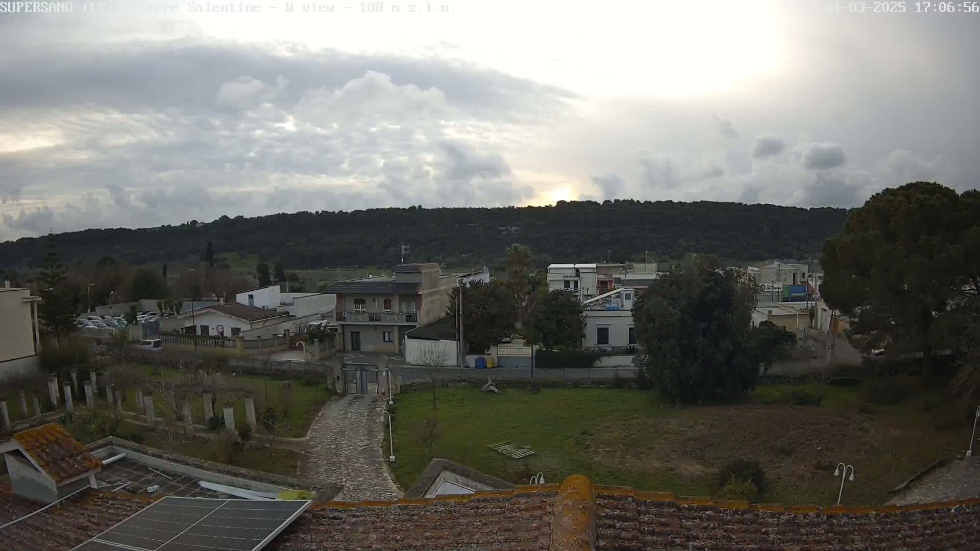 A cloudy, sunlit view of a residential area nestled against a tree-covered hill.