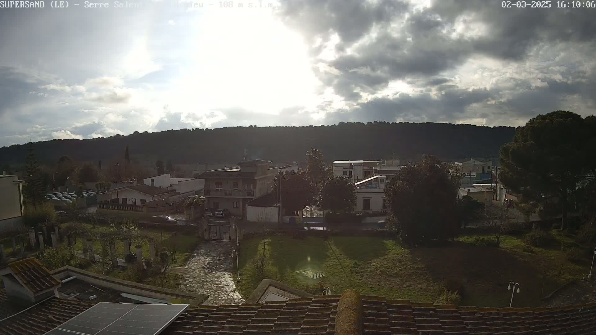 A mostly cloudy, sunlit view from a rooftop shows a small town nestled against a hillside, with a paved area and grassy lawns in the foreground.