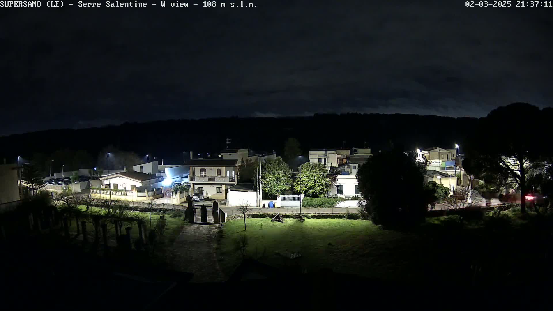 A nighttime view of a residential area under a mostly cloudy sky shows several houses illuminated by streetlights and some outdoor lights.