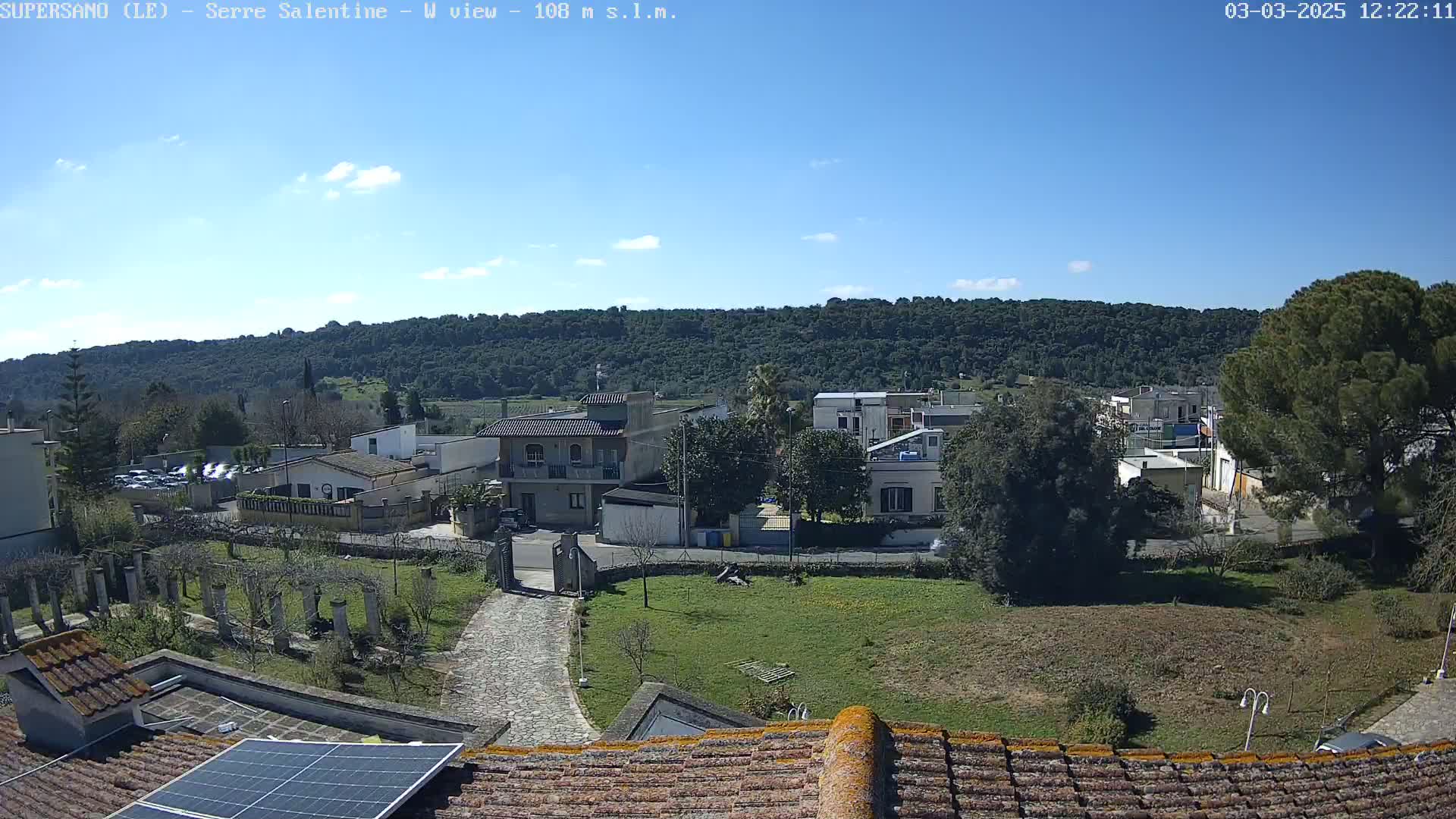 A mostly sunny view of a residential area with a hill covered in trees in the background, seen from a rooftop with a solar panel.