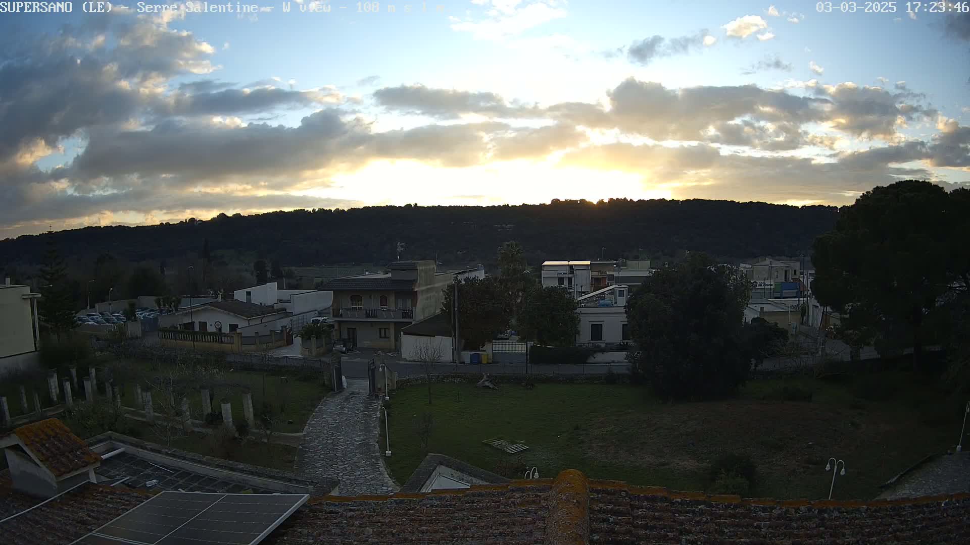 A sunset casts a warm glow over a town nestled against a hillside under a partly cloudy sky.