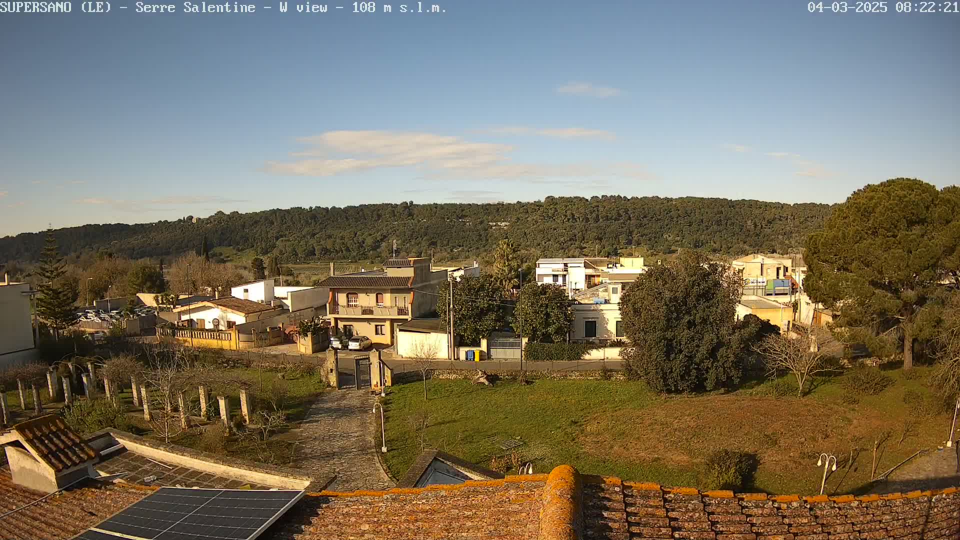 A sunny day reveals a small village nestled beside a tree-covered hill, viewed from a rooftop with solar panels.