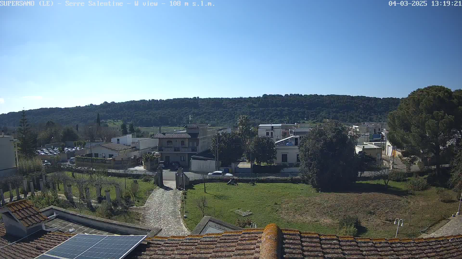 A clear, sunny day reveals a residential area nestled at the base of a tree-covered hill, viewed from a rooftop with a solar panel.