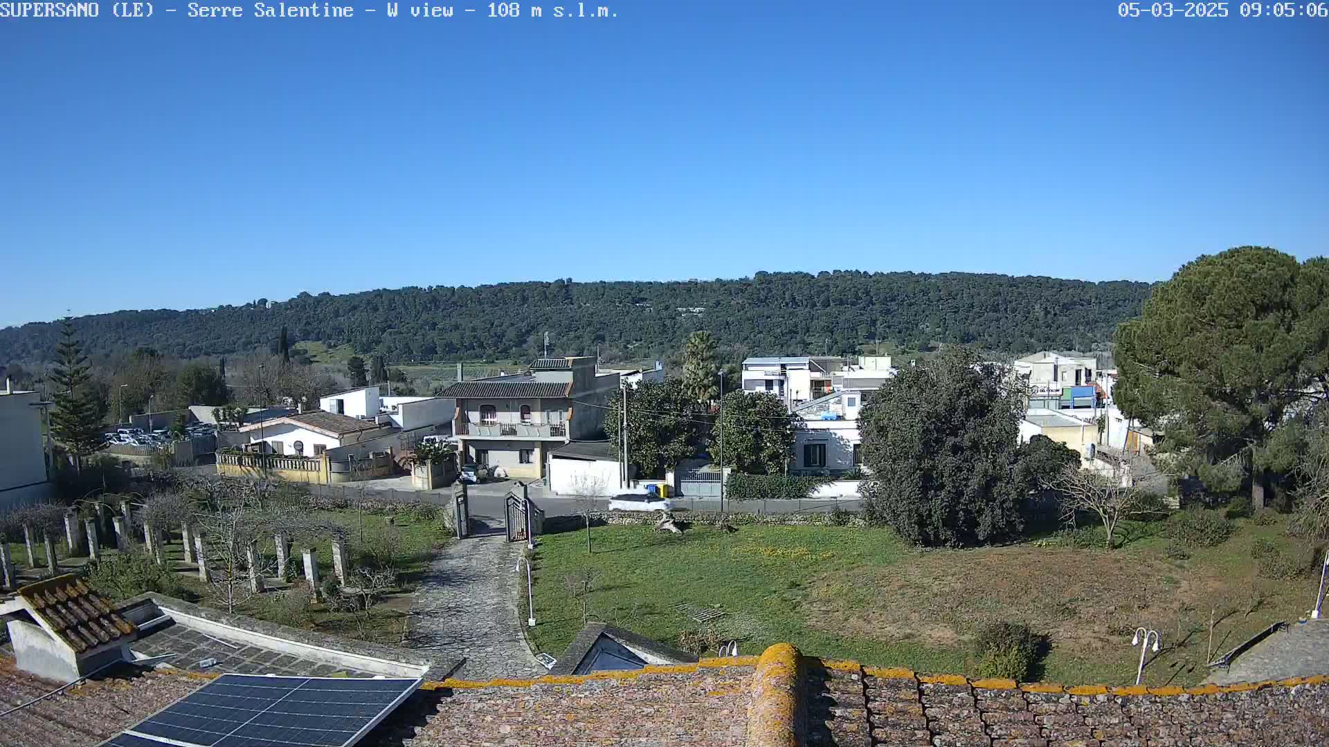 A clear, sunny day overlooking a residential area with houses nestled amongst trees and a hill in the background, viewed from a rooftop with solar panels.