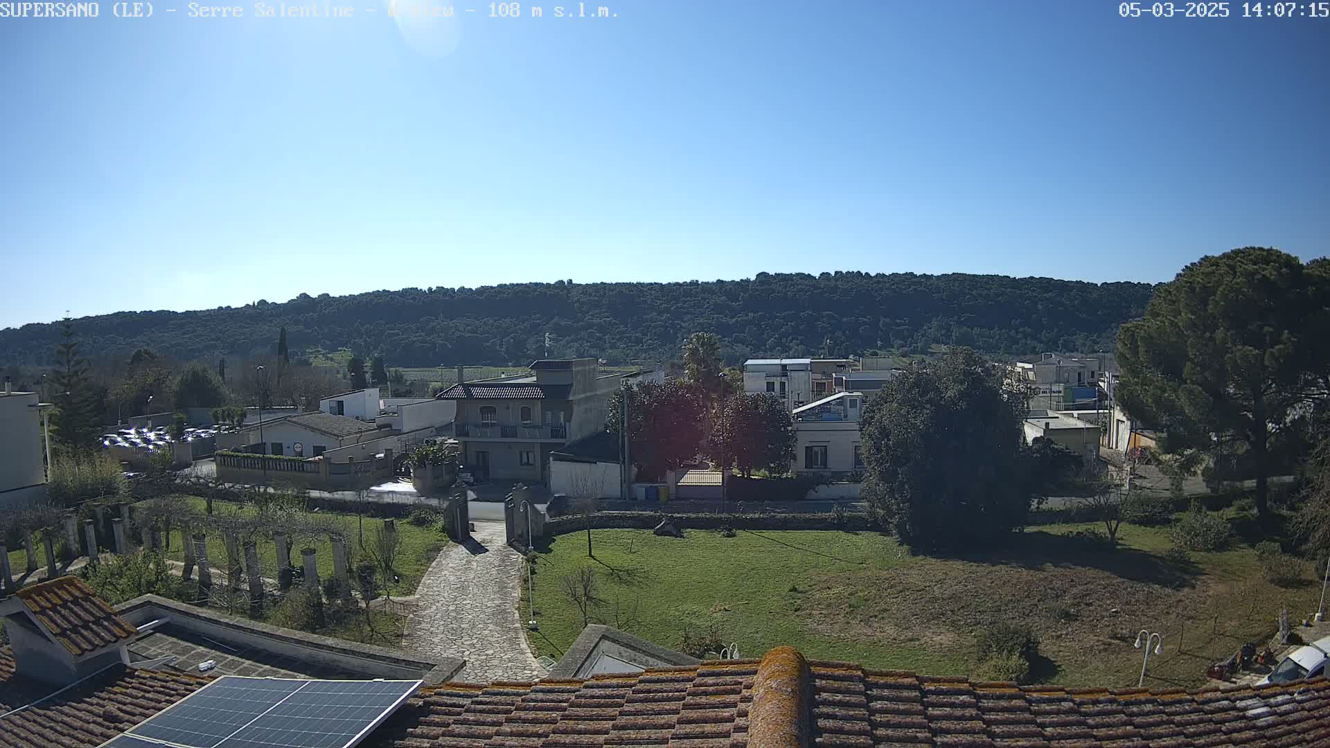 A sunny day reveals a residential area with low buildings and a paved path leading to a gated entrance, set against a backdrop of a large, forested hill.