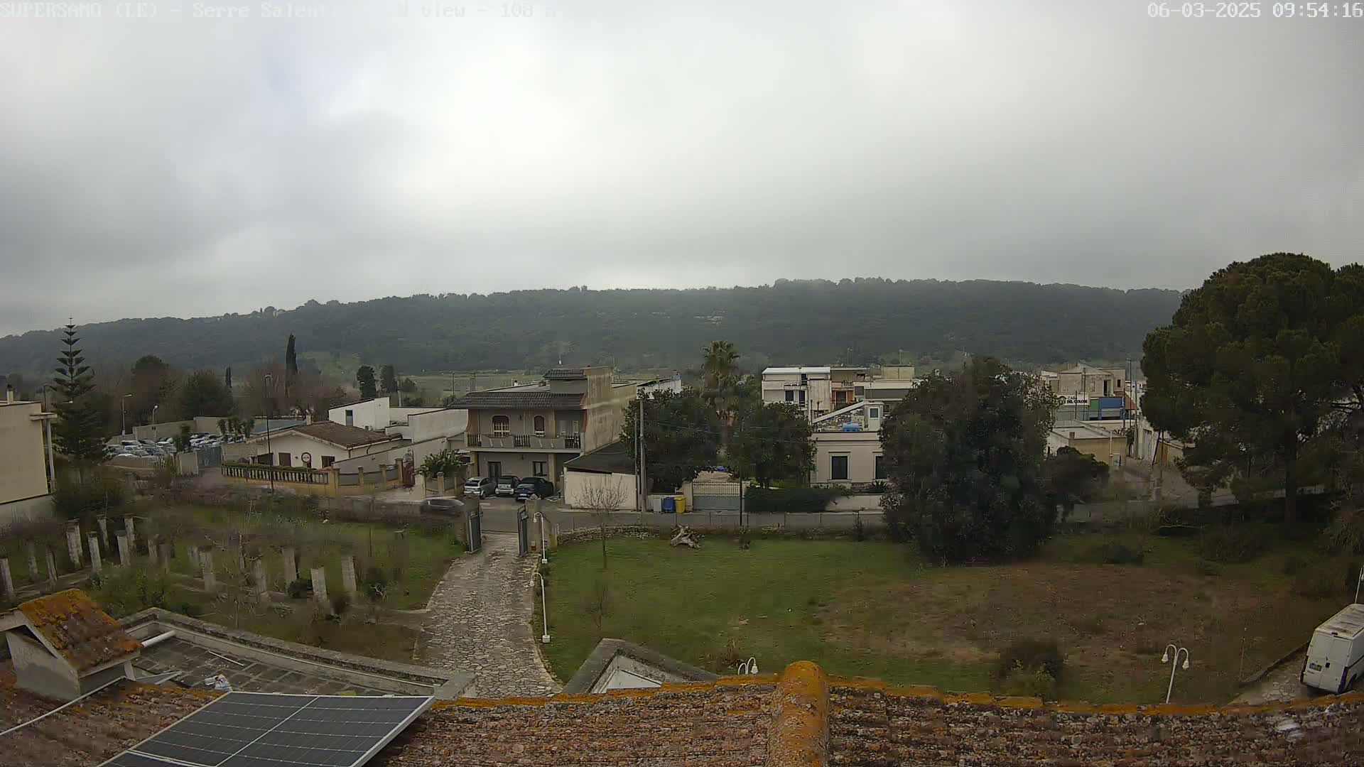 A view of a residential area with low buildings and a tree-covered hill in the background on an overcast day.