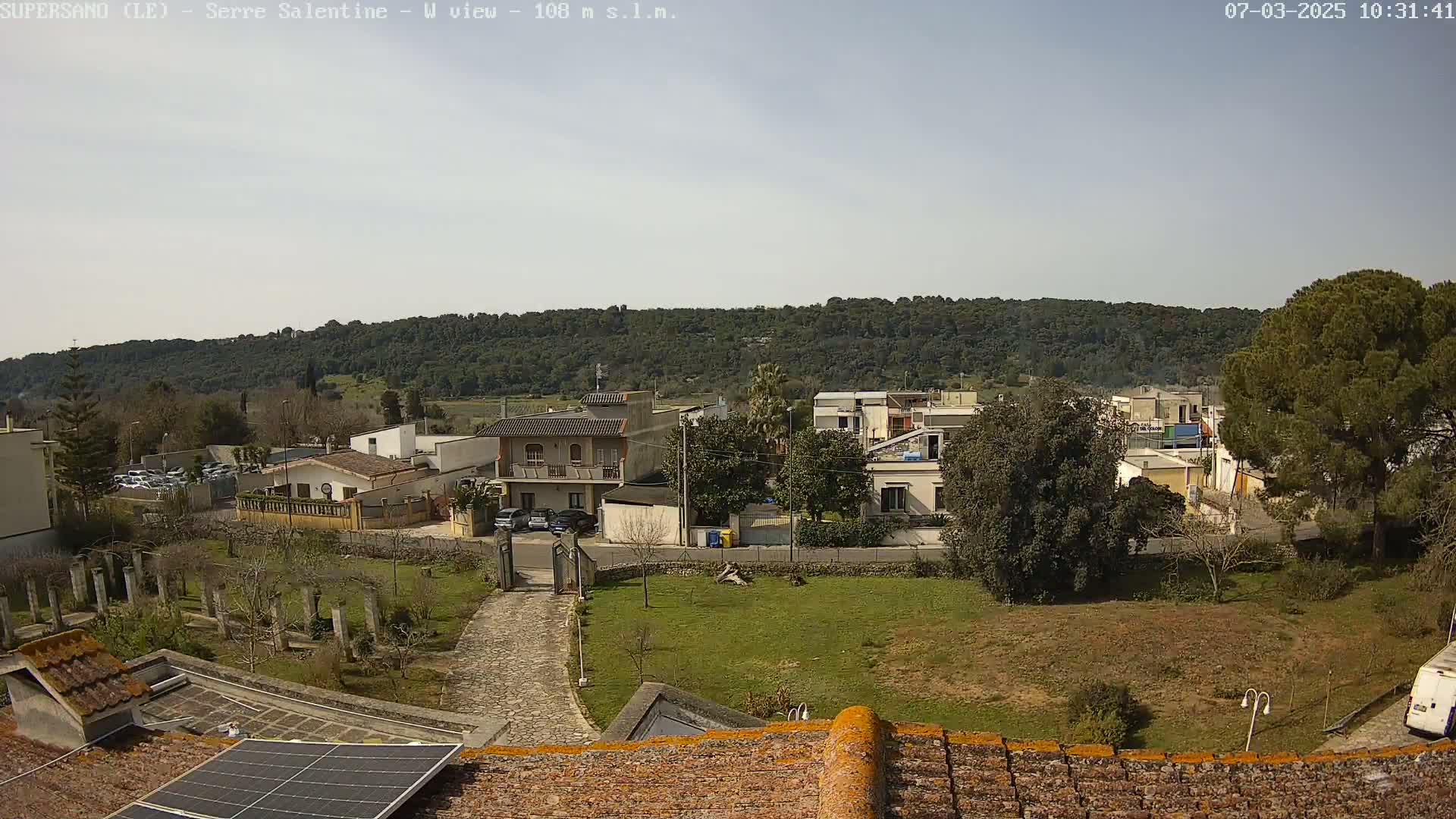 A residential area with low buildings and a hill covered in trees in the background under a mostly clear sky.