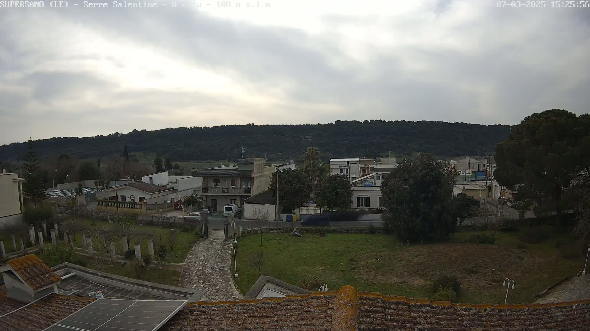 A cloudy day overlooks a residential area with low buildings and green spaces, nestled against a backdrop of a verdant hill.