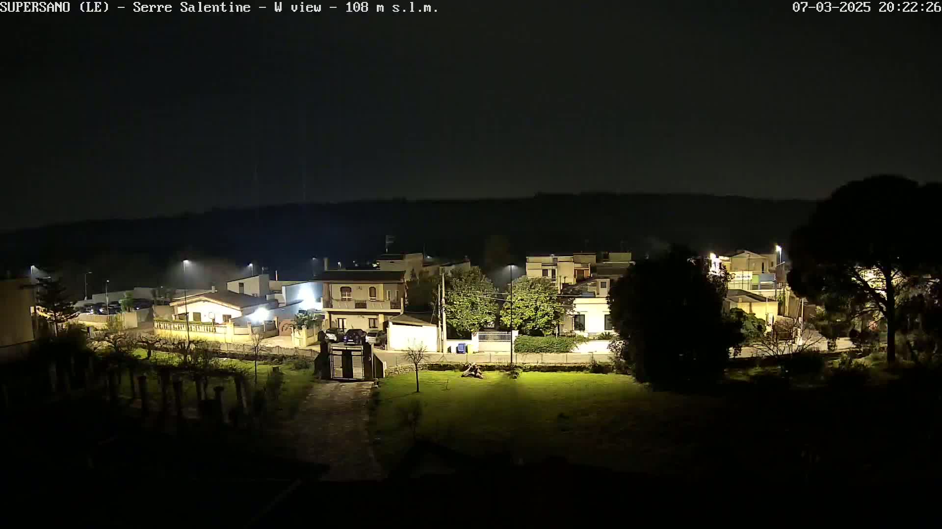A nighttime, illuminated view of a residential area with houses, trees, and a grassy lawn under a dark, clear sky.
