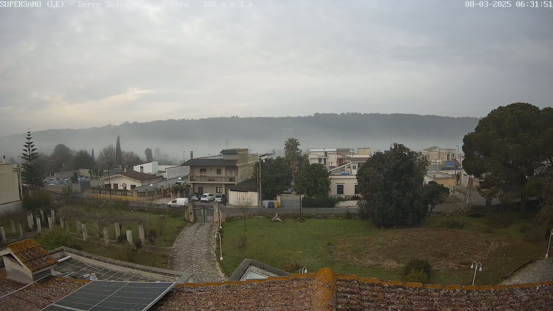 A foggy, overcast day reveals a residential area nestled against a low-lying, fog-covered hillside, with a paved path and a solar panel visible in the foreground.