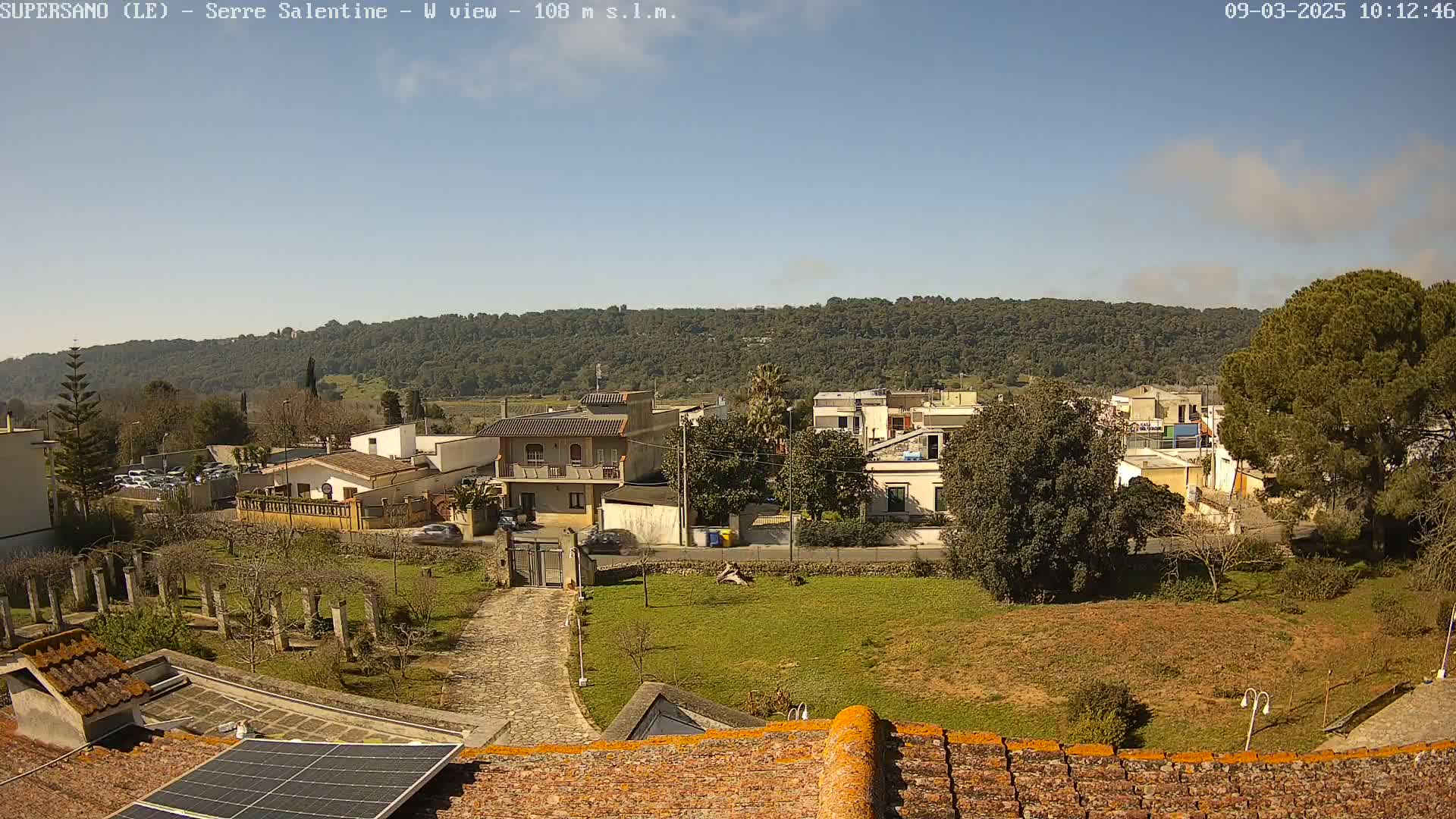 A view from a rooftop shows a sunny day overlooking a collection of low buildings nestled amongst green vegetation, with a hill covered in trees in the background.