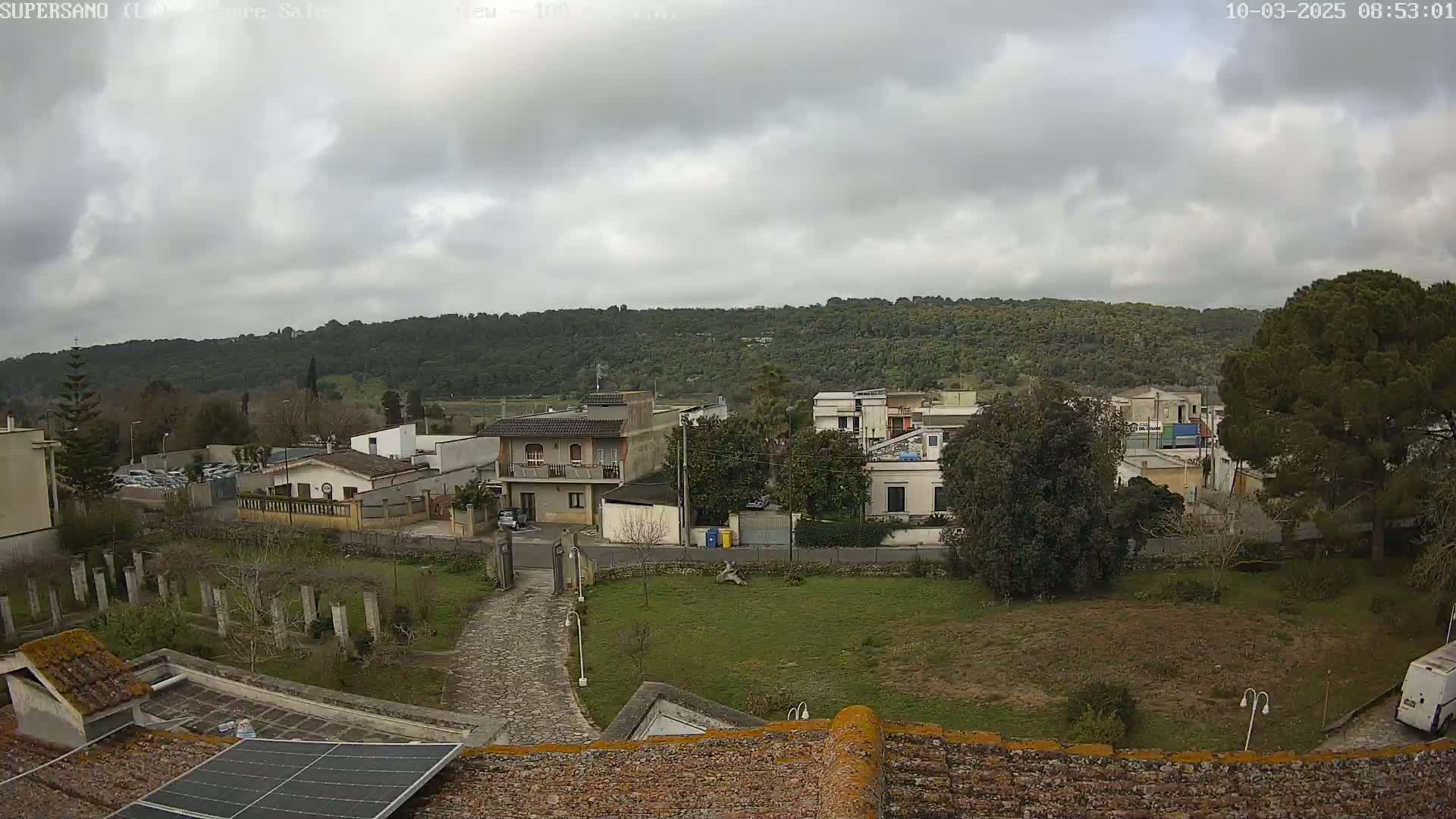 An overcast day reveals a residential area nestled at the foot of a verdant hill, viewed from a rooftop with a solar panel.