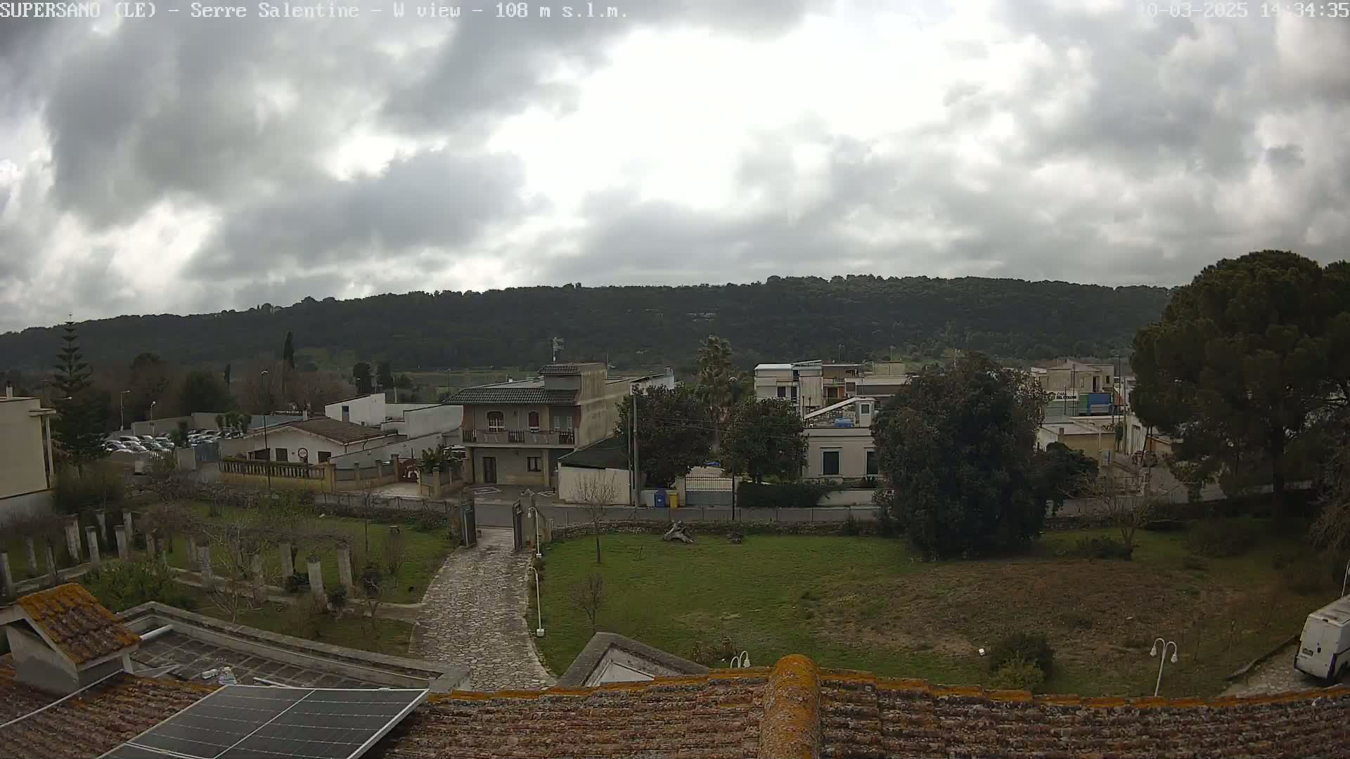 A cloudy day reveals a residential area nestled beneath a tree-covered hill, with a paved pathway leading to a building featuring solar panels on its roof.
