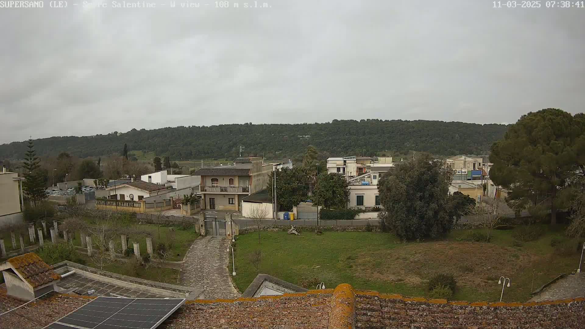 A residential area with low buildings and green spaces is visible under an overcast sky, nestled below a line of green hills in the distance.