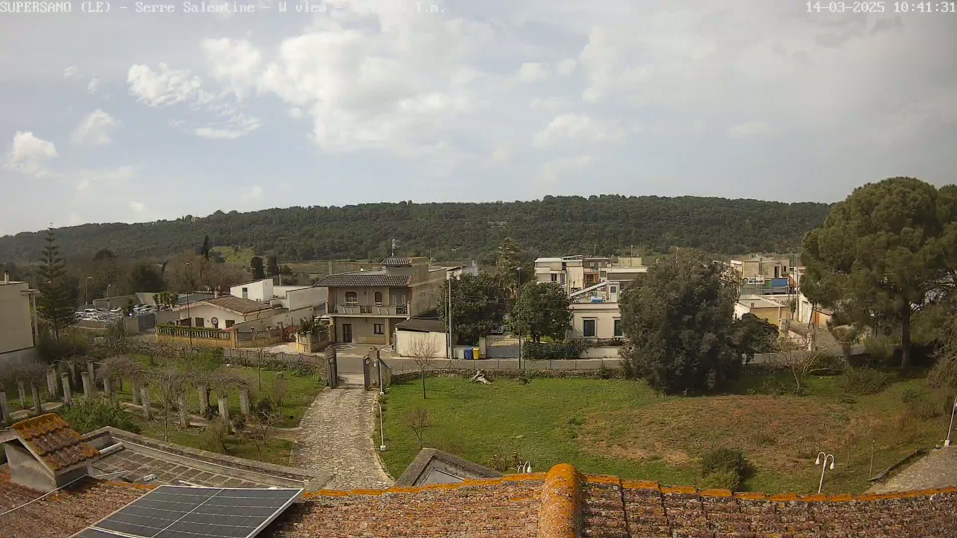 A partly cloudy day reveals a view of a residential area nestled against a tree-covered hillside, seen from a rooftop with a solar panel.
