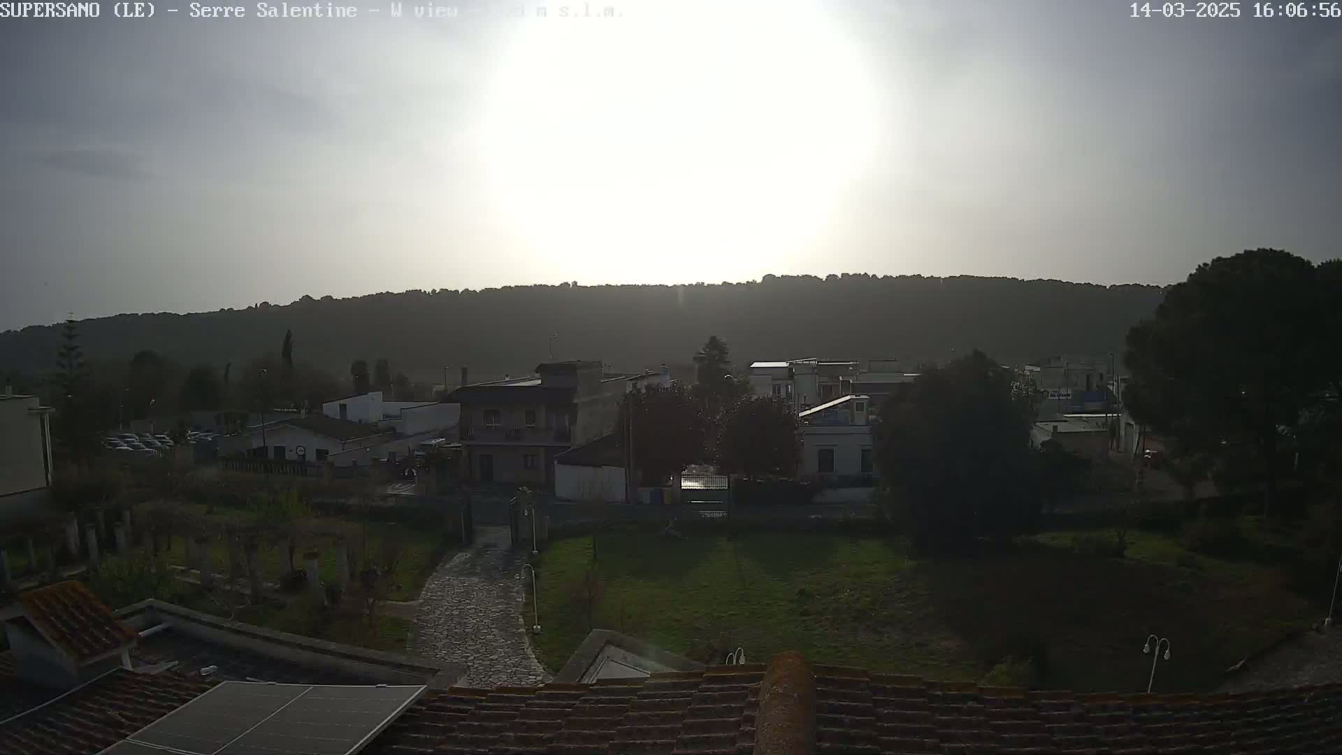 A bright, hazy sun shines over a residential area nestled against a tree-covered hillside.