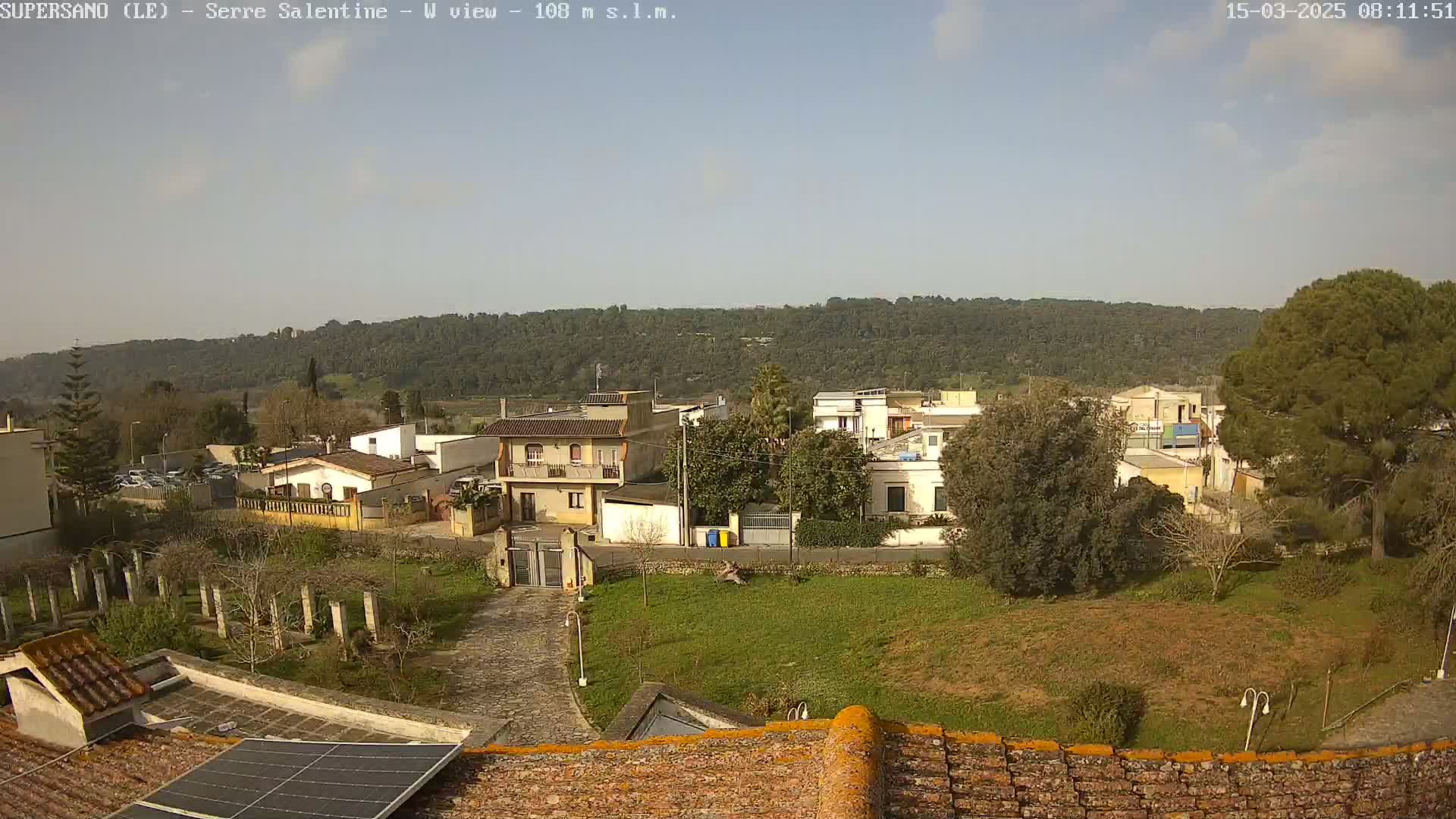 A mostly sunny day reveals a residential area nestled against a hilly, wooded landscape, viewed from a rooftop with solar panels.