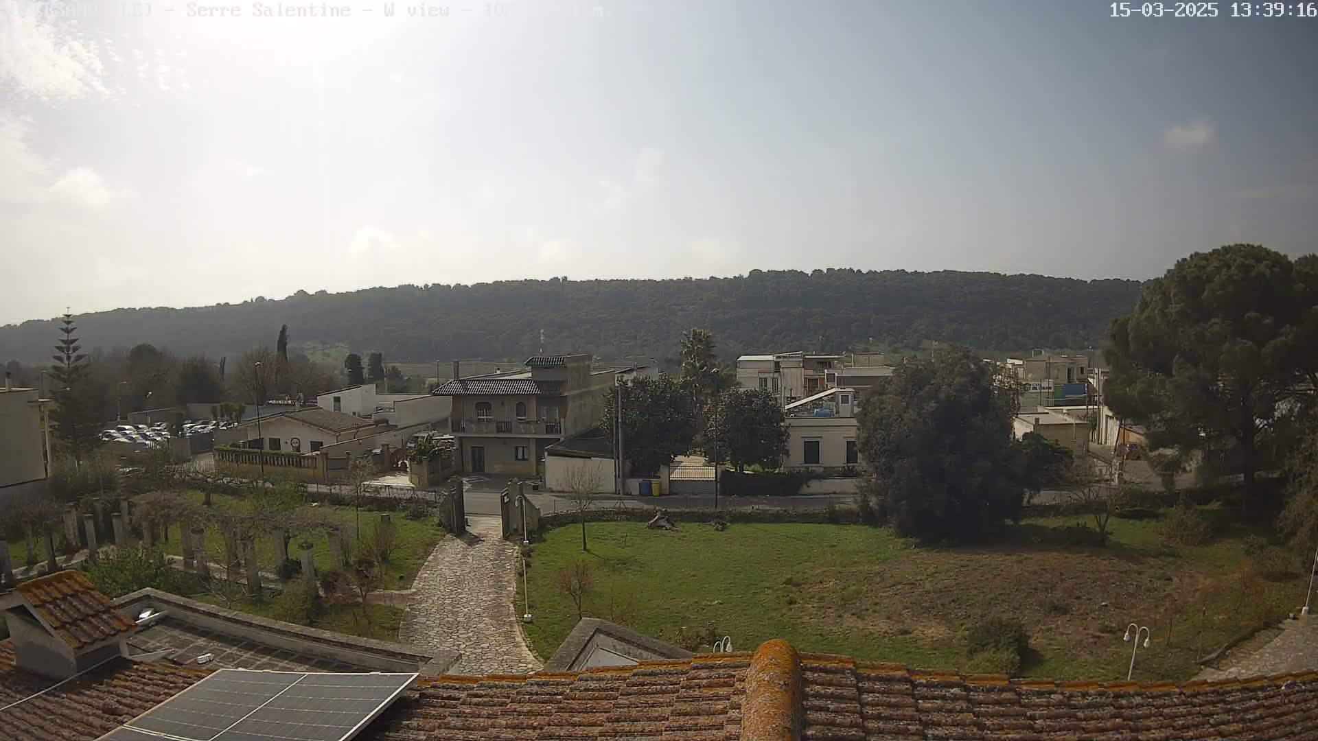A sunlit view from a rooftop shows a residential area with low buildings and green lawns, all set against a backdrop of a tree-covered hill.