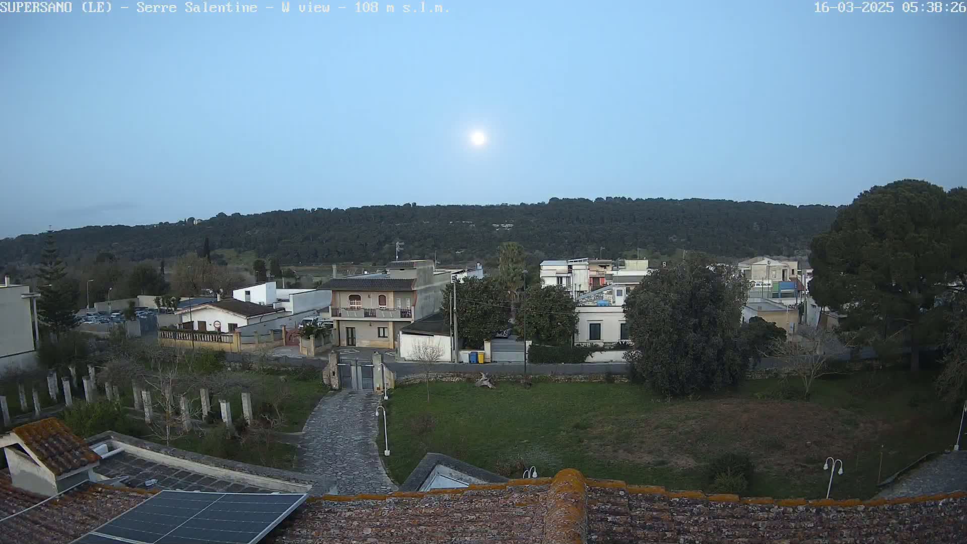 A sunlit, clear sky overlooks a residential area with low-lying buildings and a hill covered in trees in the background.