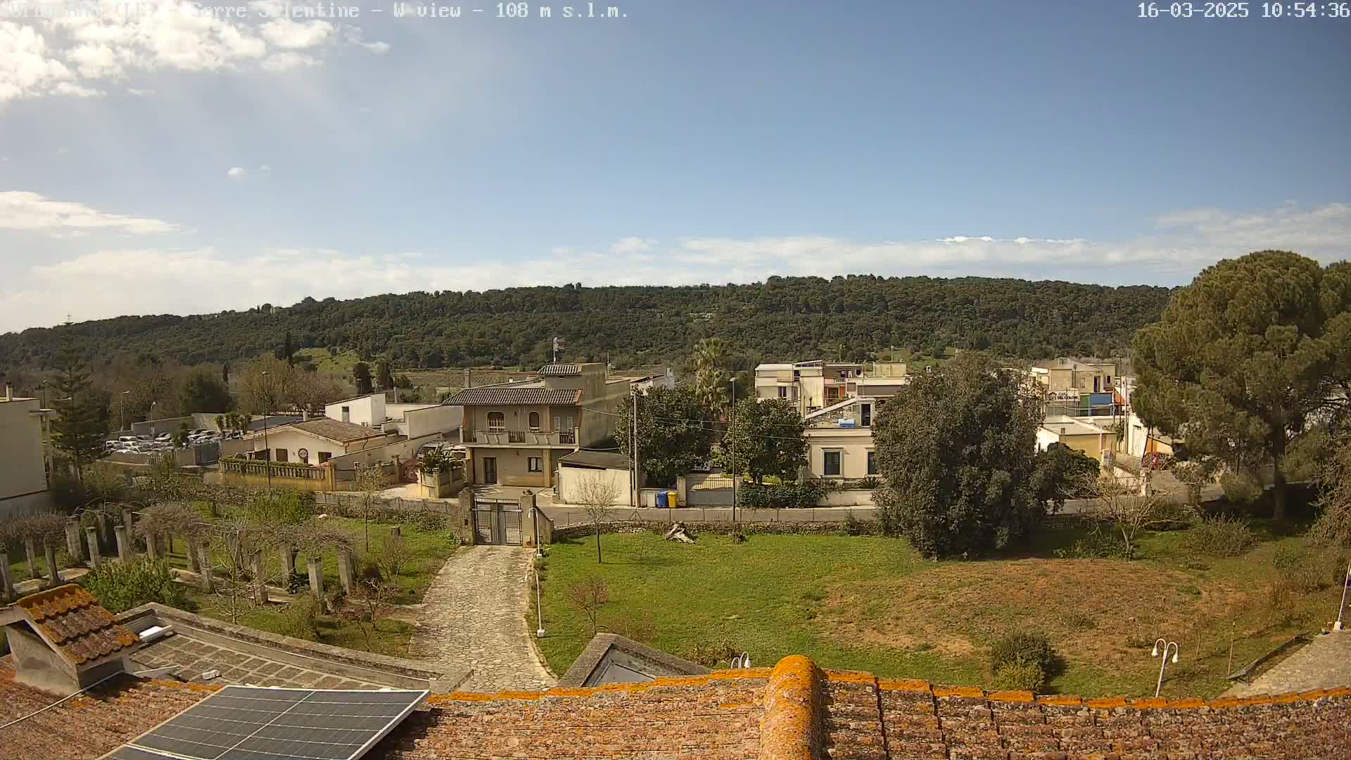 A partly sunny day reveals a residential area nestled at the base of a tree-covered hill, viewed from a rooftop with a solar panel.