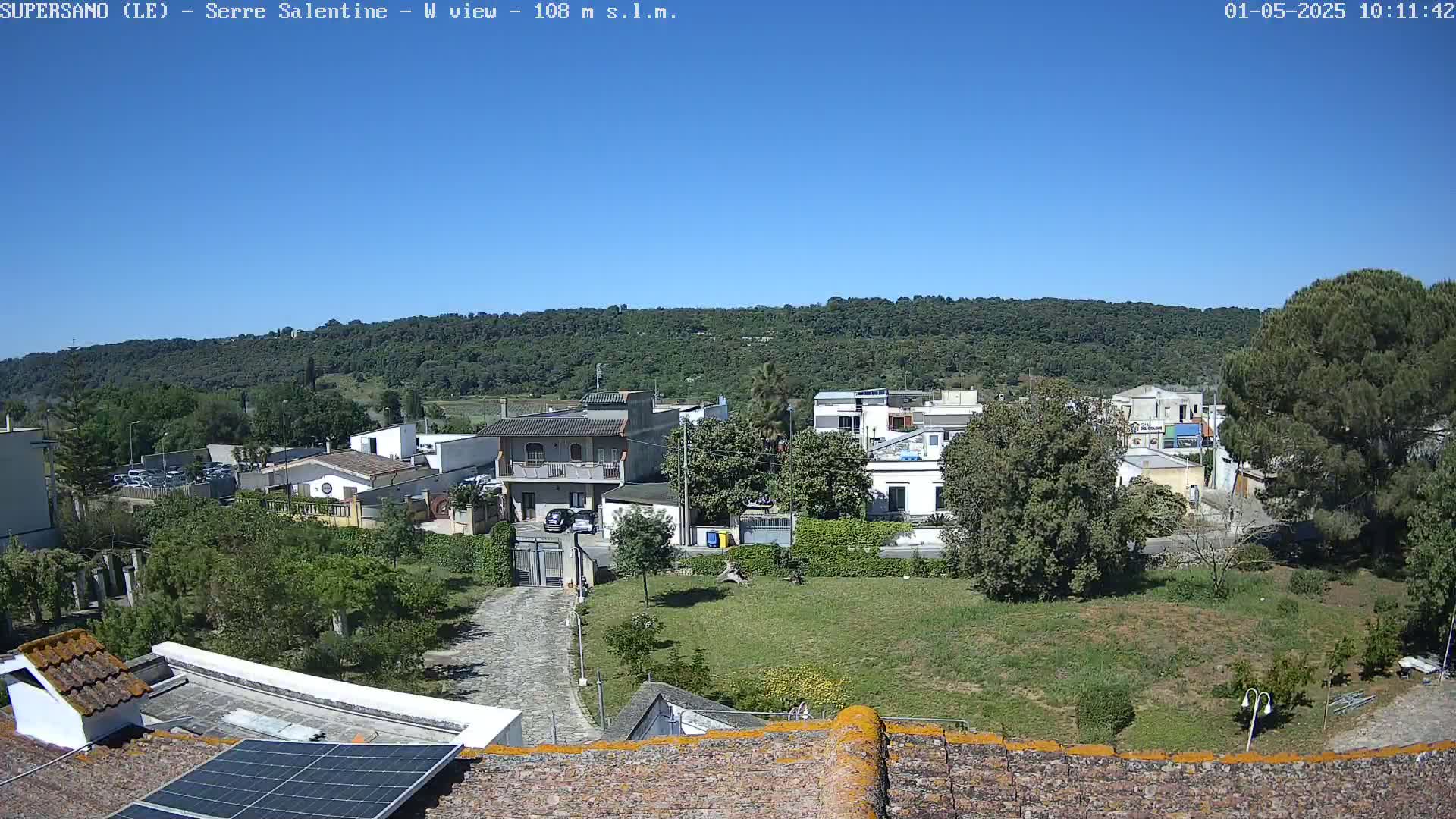 A clear, sunny day reveals a view of a residential area nestled against a lush, green hillside, seen from a rooftop with a solar panel.