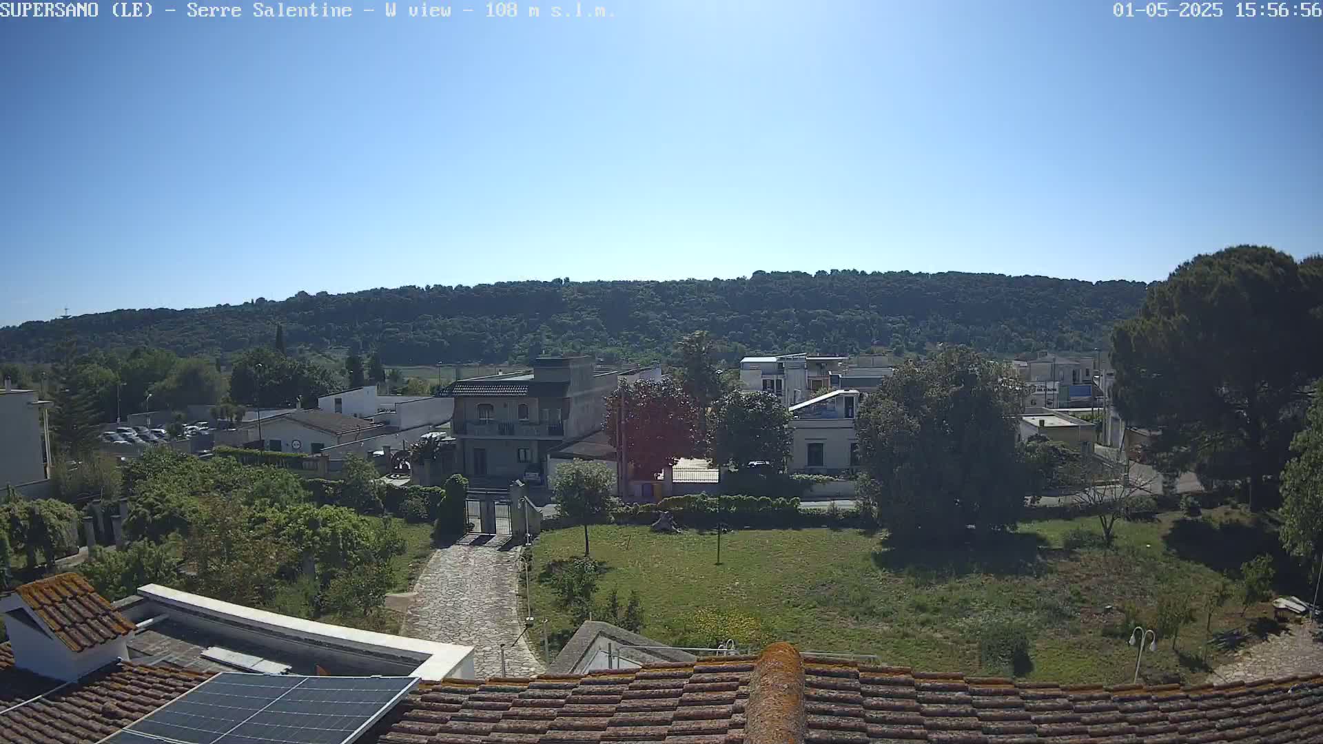 A clear, sunny day reveals a view of buildings and lush green vegetation, nestled beneath a tree-covered hill.