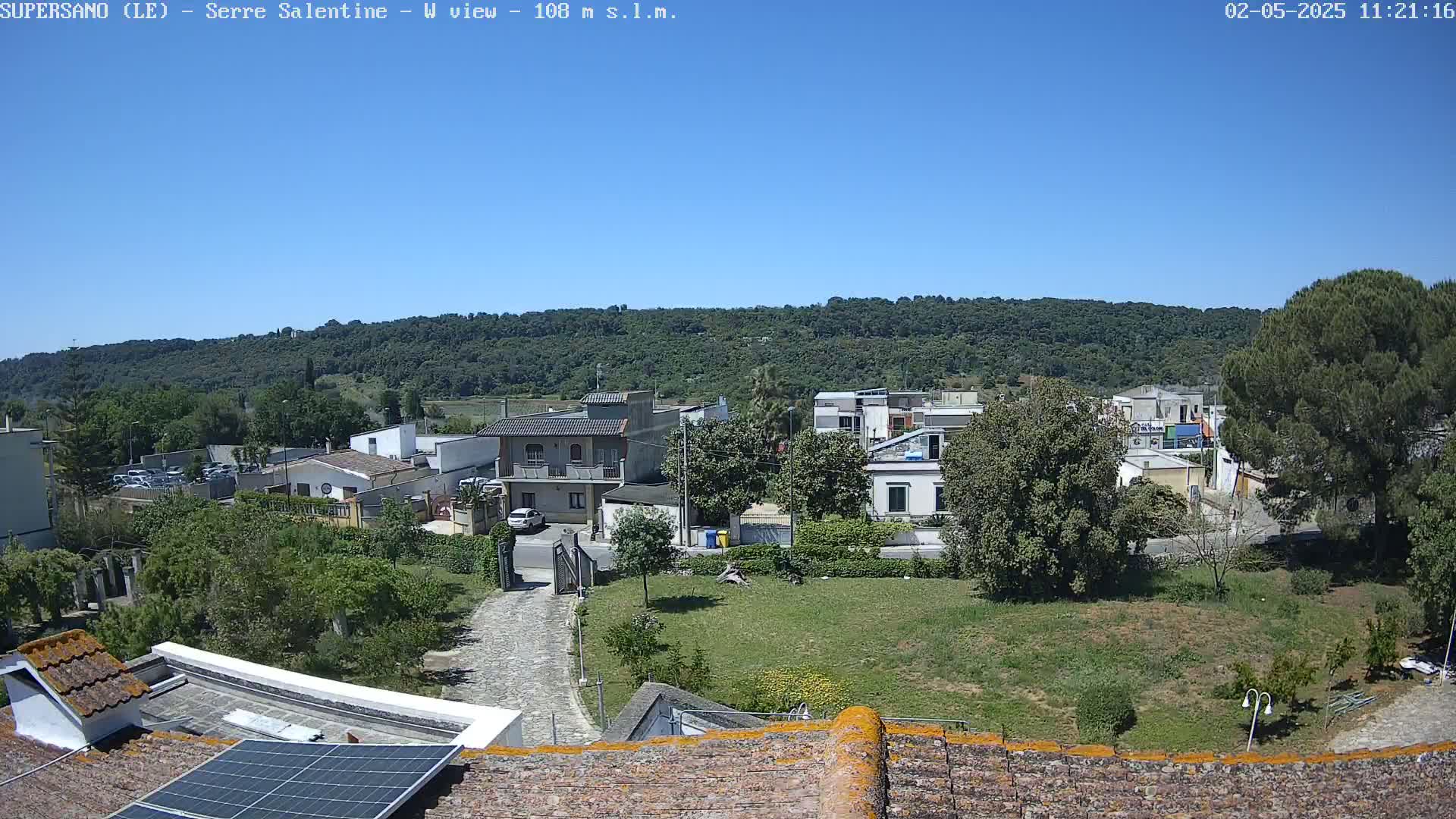 A sunny, high-angle view of a residential area nestled against a verdant hillside, featuring low buildings, trees, and a paved driveway.