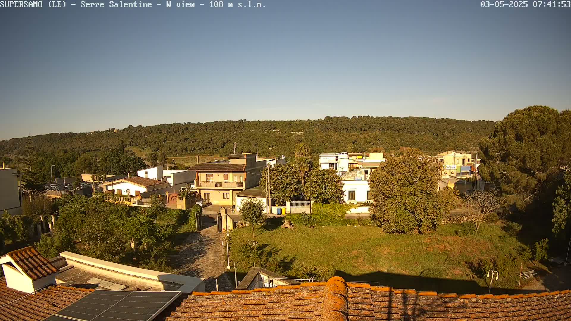 A sunlit village with low buildings and green vegetation is nestled below a hill covered in dark green trees under a clear sky.