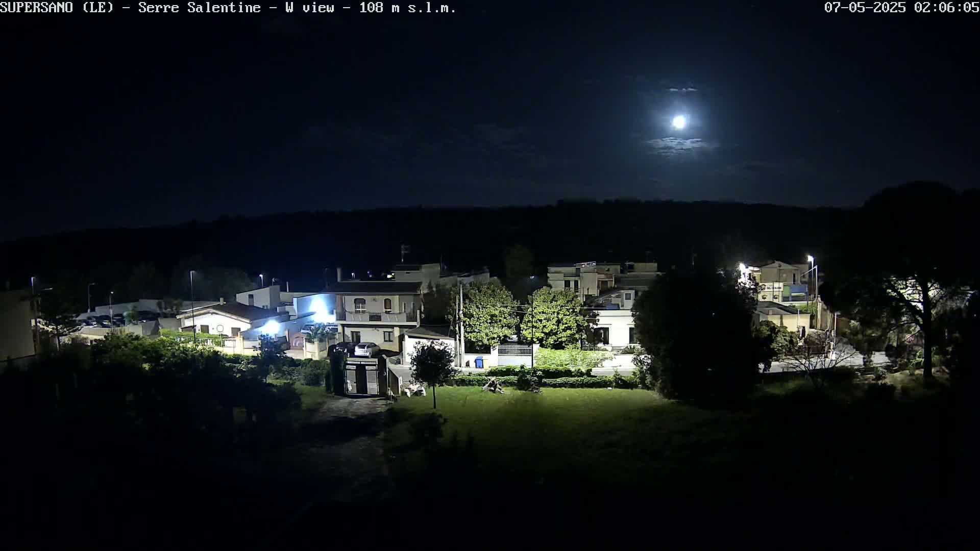 A brightly lit moon illuminates a residential area at night, with houses and trees visible under a mostly clear sky.