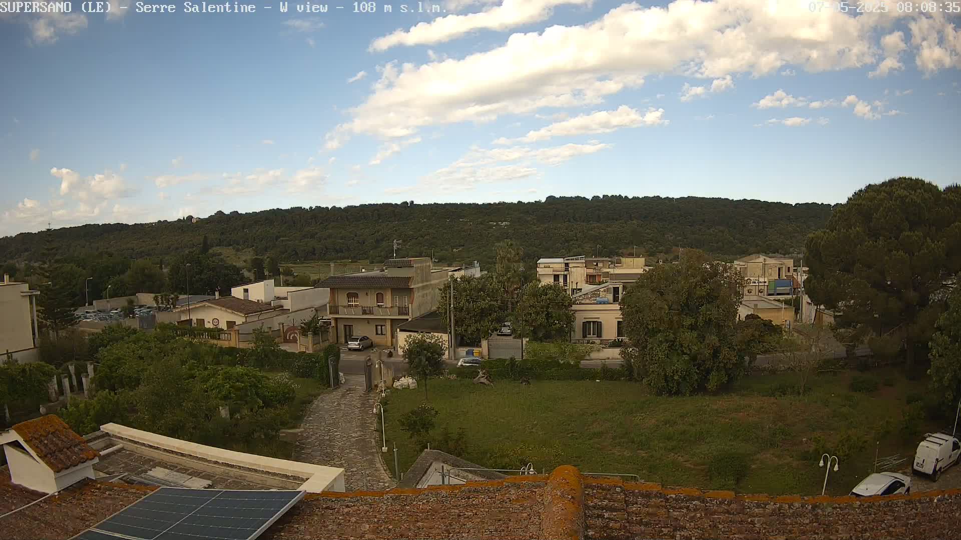 A partly cloudy sky overlooks a residential area with low-lying buildings, green lawns, and a tree-covered hill in the background.