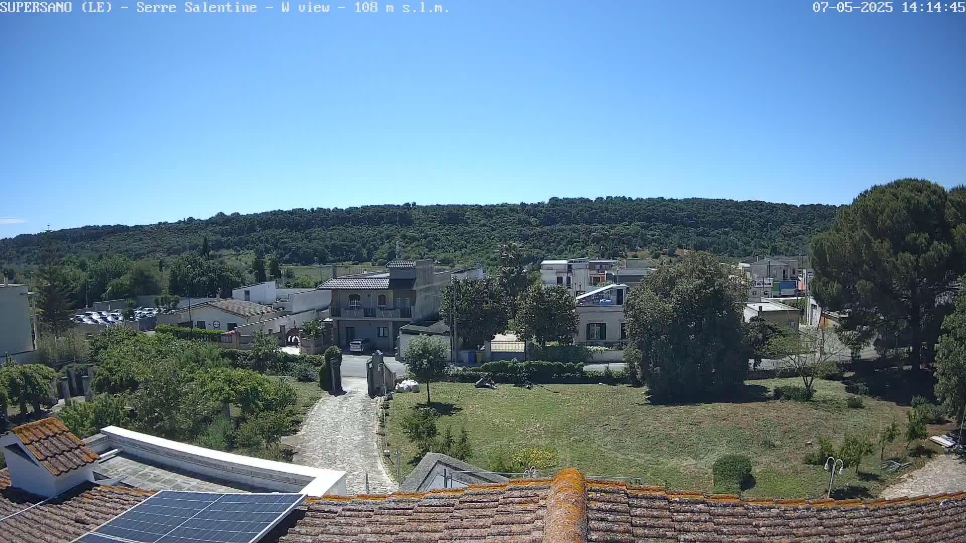 A sunlit view of a small town nestled amongst green hills under a clear blue sky, seen from a rooftop with solar panels.