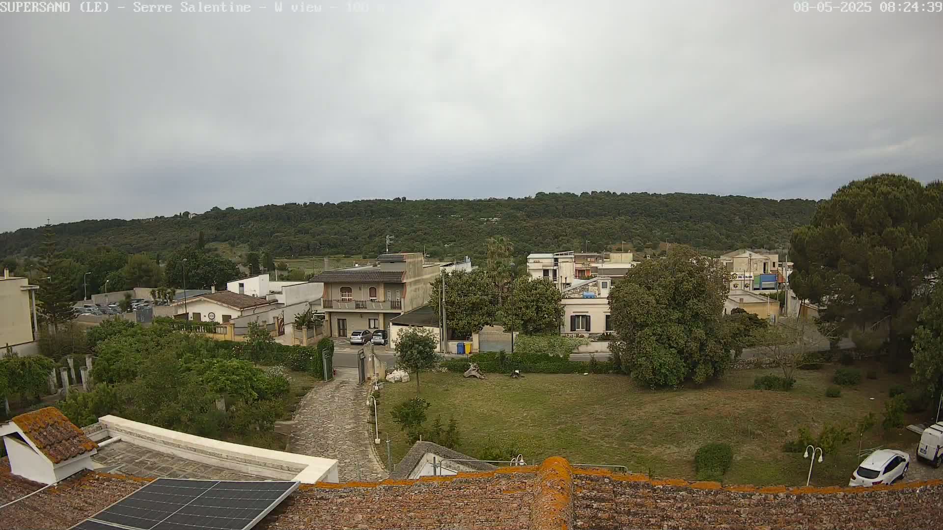 A view from a rooftop shows a residential area with overcast skies, green hills in the background, and a paved driveway leading to houses.