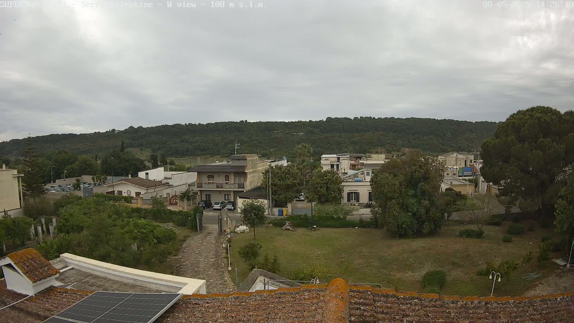 A view from a rooftop shows a residential area with low buildings and green spaces under an overcast sky.