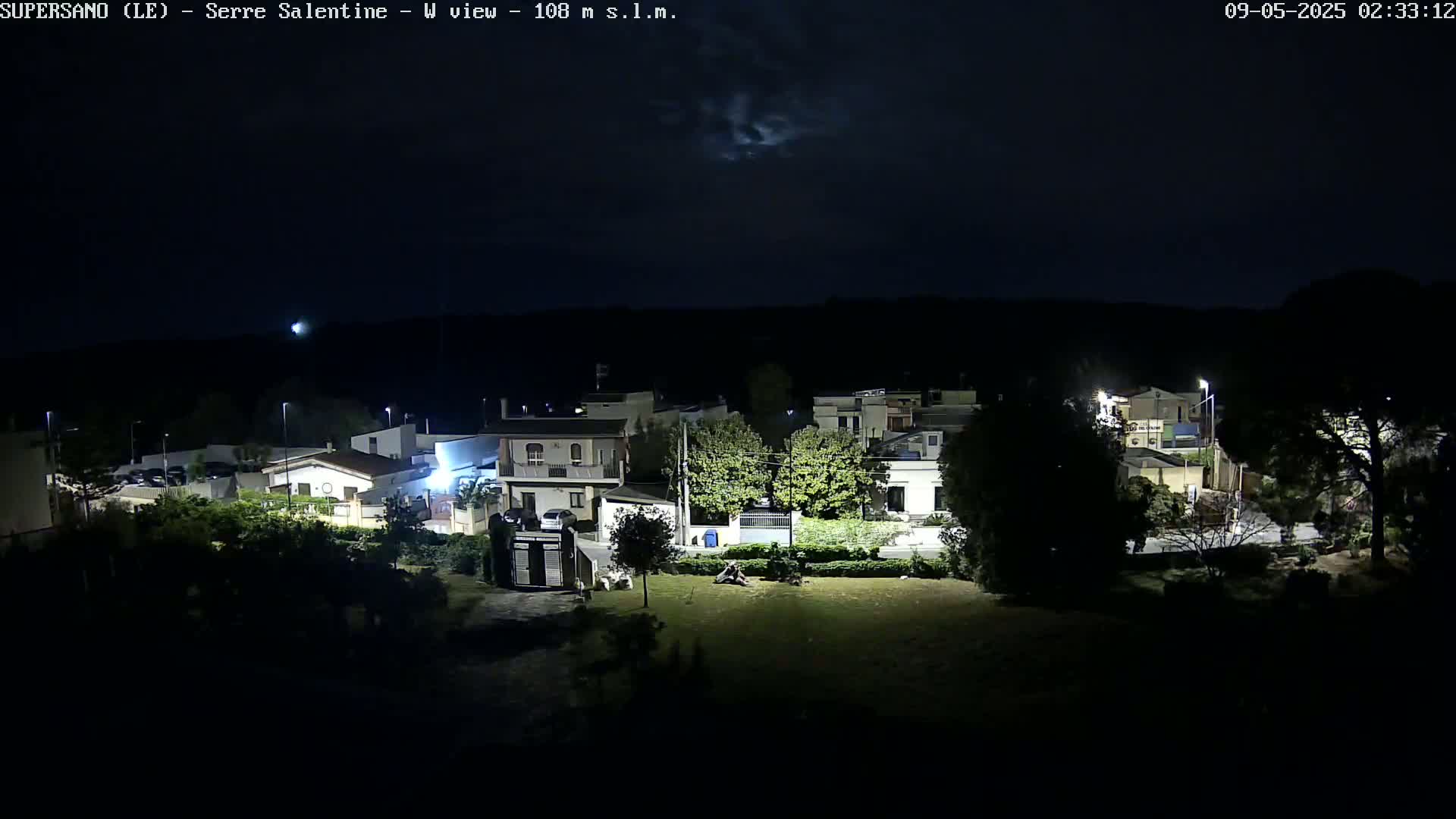 A nighttime view of a residential area, under a mostly clear sky, shows several houses with streetlights illuminating the scene.