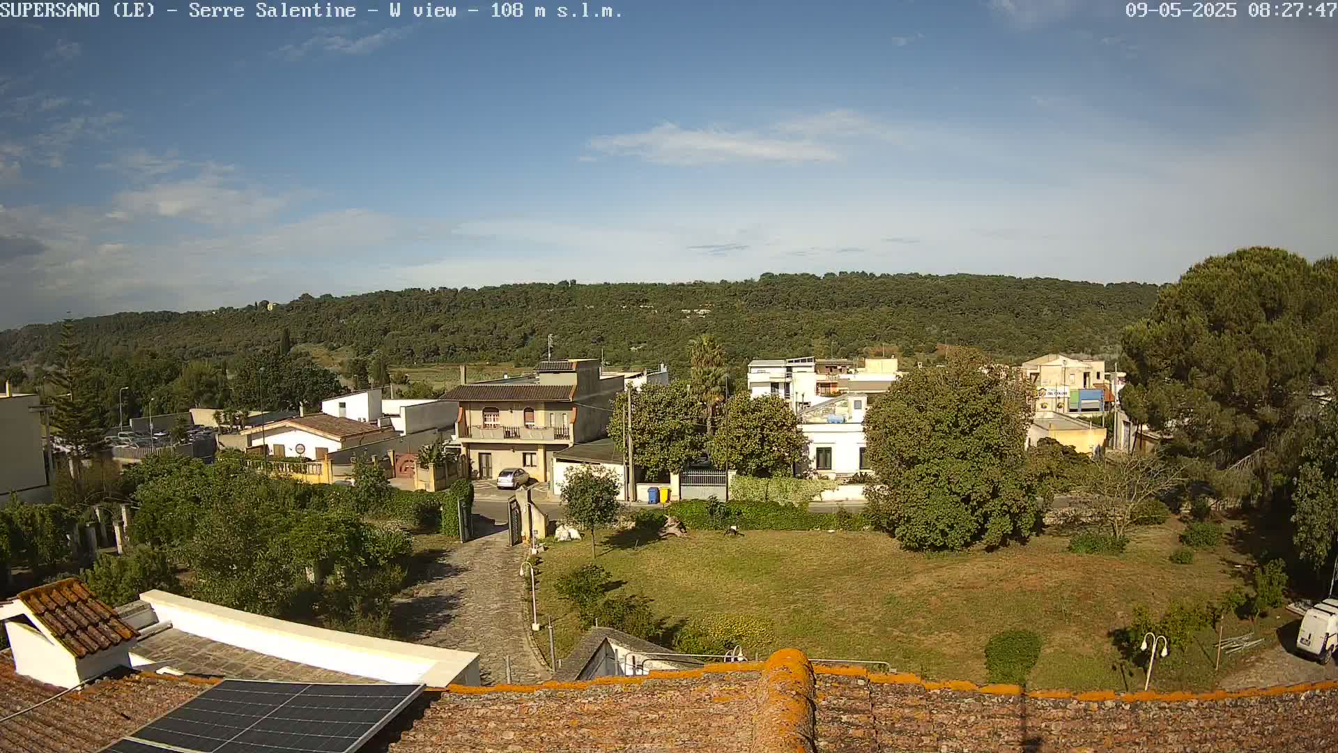 A mostly sunny day reveals a residential area with low buildings and green spaces, backed by a hill covered in dark green vegetation.